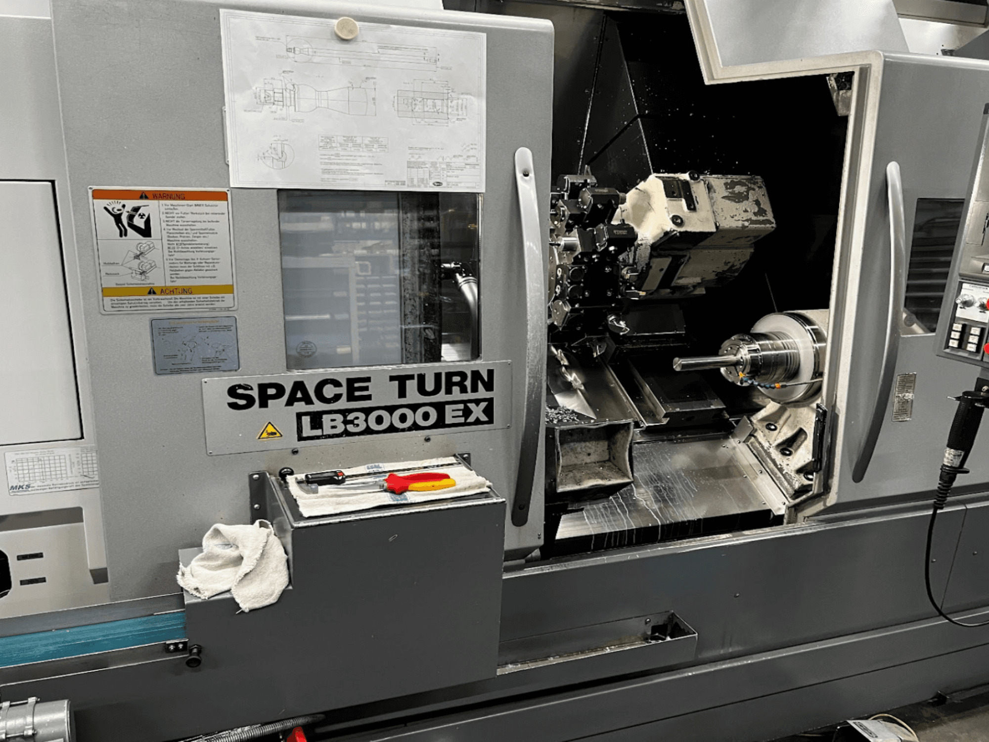 Горизонтальный токарный станок Okuma LB3000EX MYW, вид спереди, видны панель управления, инструменты и детали рабочего пространства.