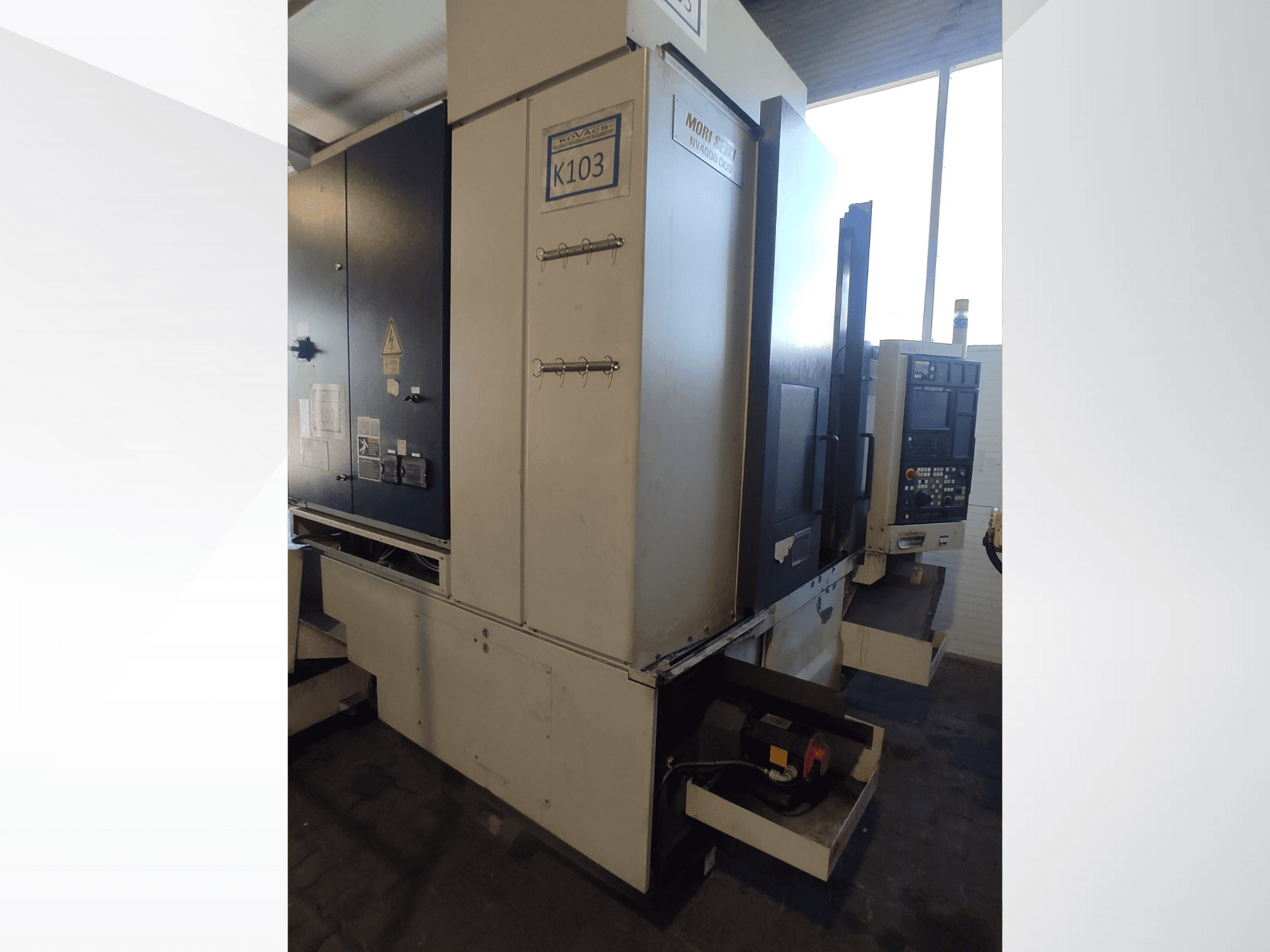 Станок с ЧПУ Mori Seiki K103 с видом спереди, видны панель управления, наклейки безопасности и электрические компоненты.