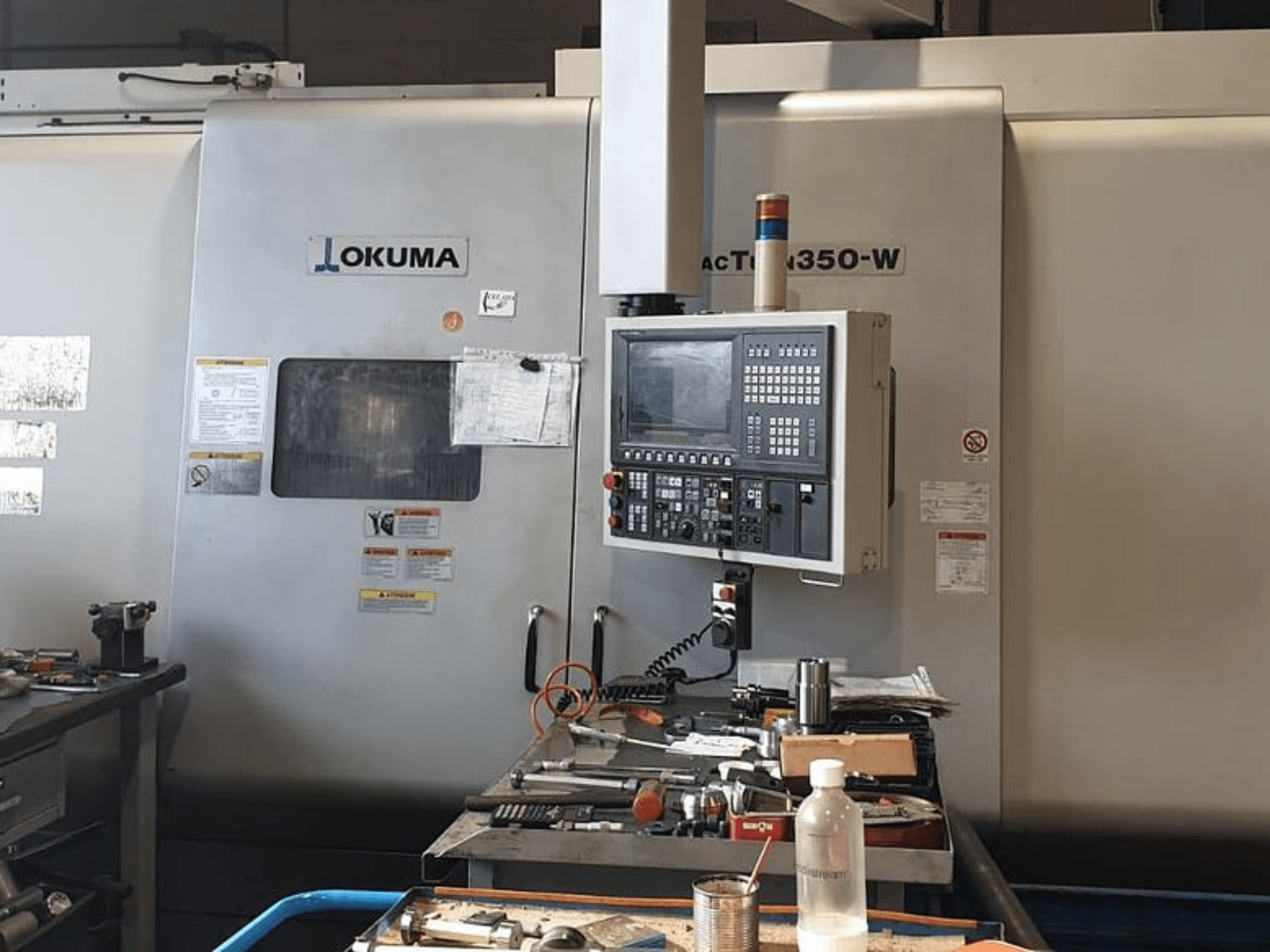 Токарно-фрезерный центр Okuma MAC TURN 350-W, вид спереди с панелью управления и рабочей зоной, загроможденной инструментами и бутылкой.