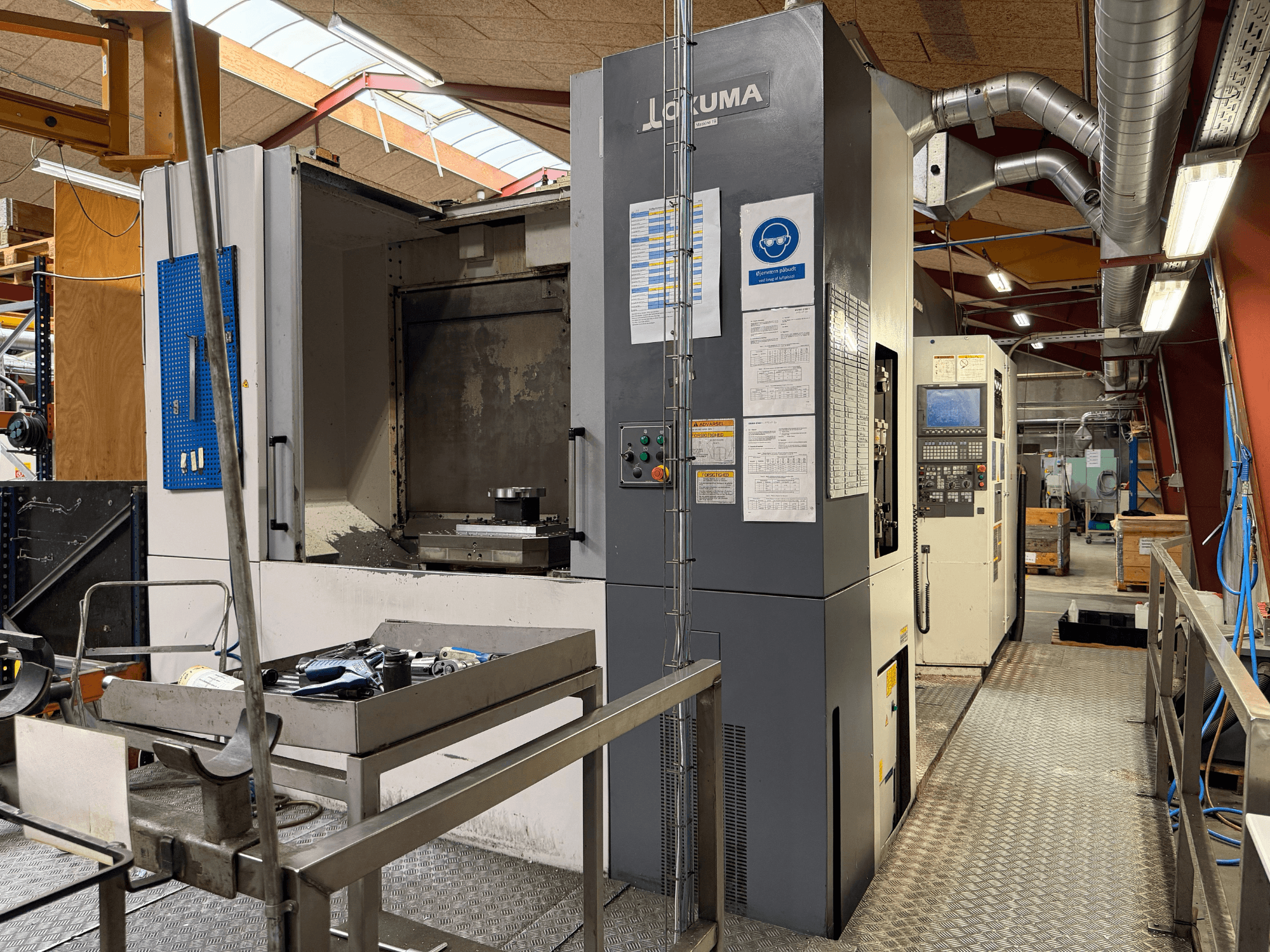 Горизонтальный обрабатывающий центр Okuma MA-600HB, вид спереди, на котором видны инструменты на металлическом столе и органы управления станком.