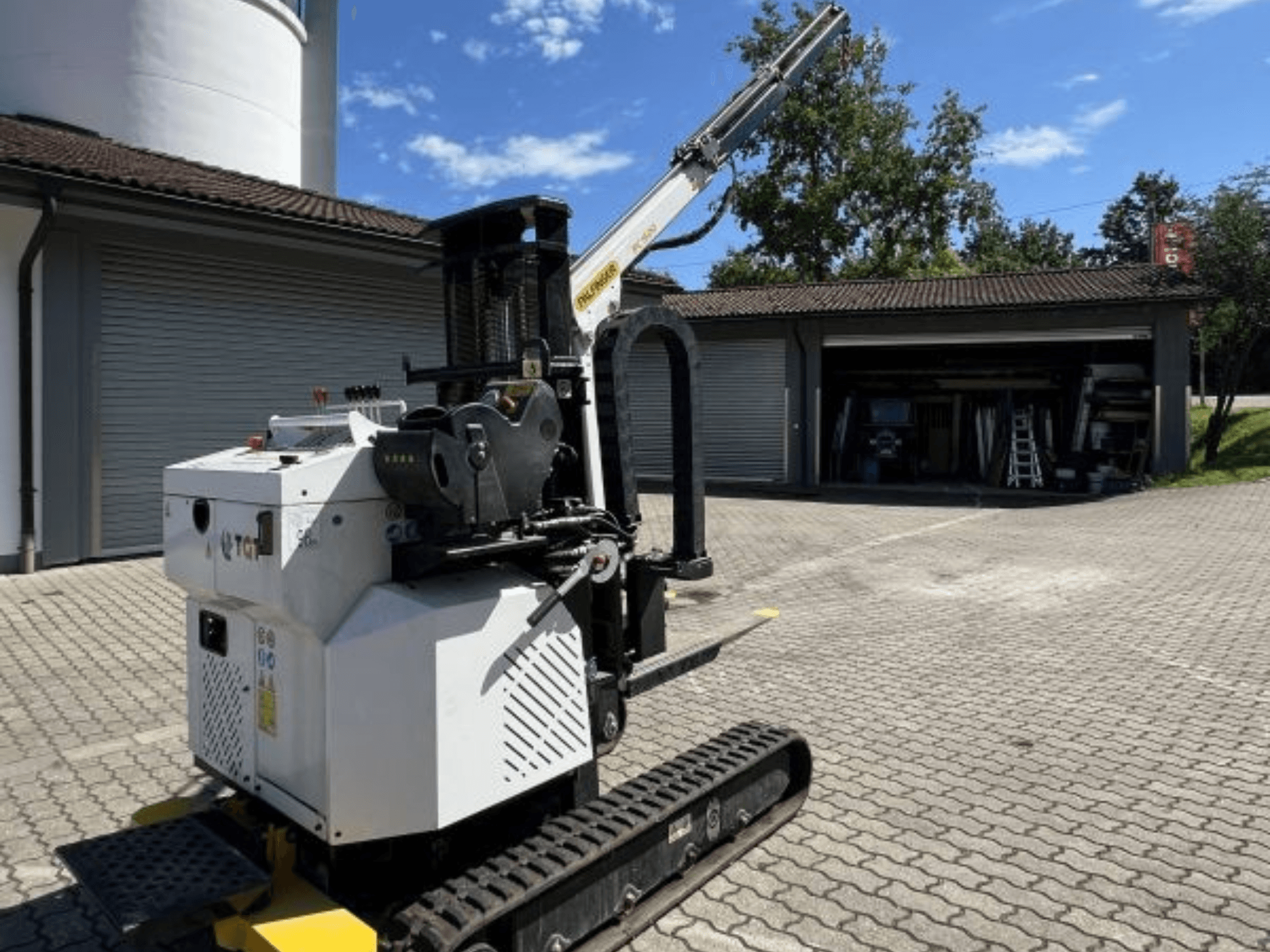 Строительный мини-кран TGI, вид сбоку, видны рельсы и установленная стрела, припаркован на кирпичной площадке у гаража.