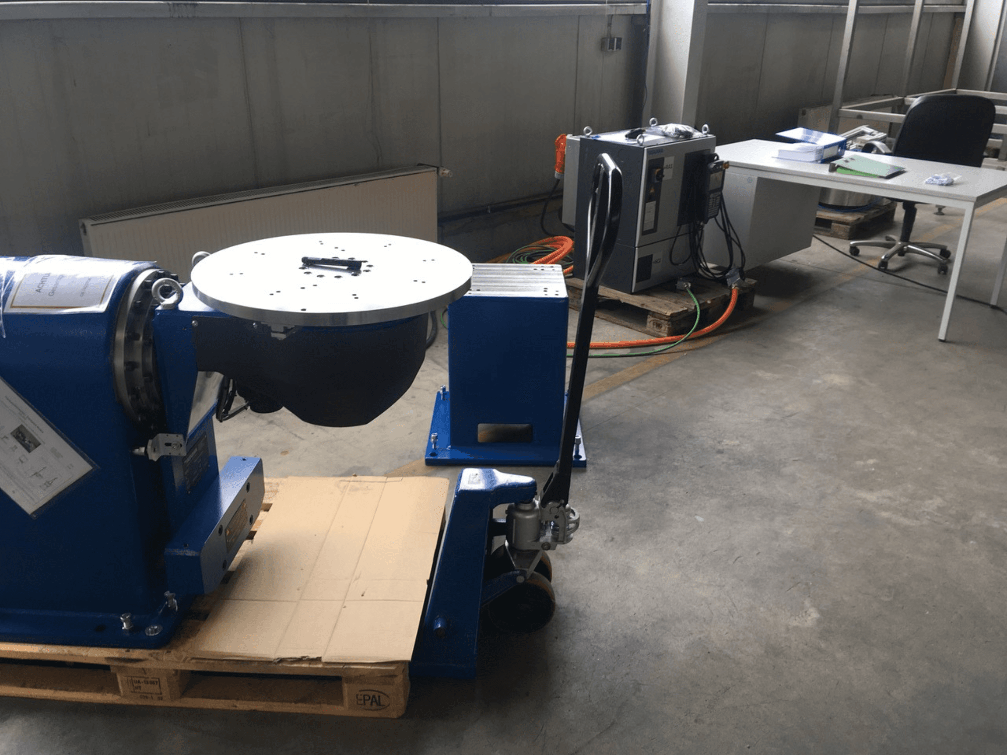 Синее промышленное оборудование с круглым основанием и панелью управления, вид спереди, на деревянном поддоне в офисном помещении.