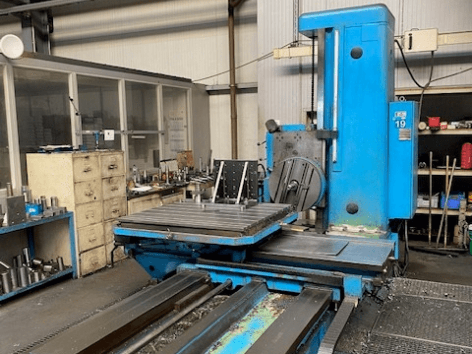 Синий фрезерный станок с видом спереди, модель № 19, на полу мастерской с инструментами и шкафом для хранения на заднем плане.