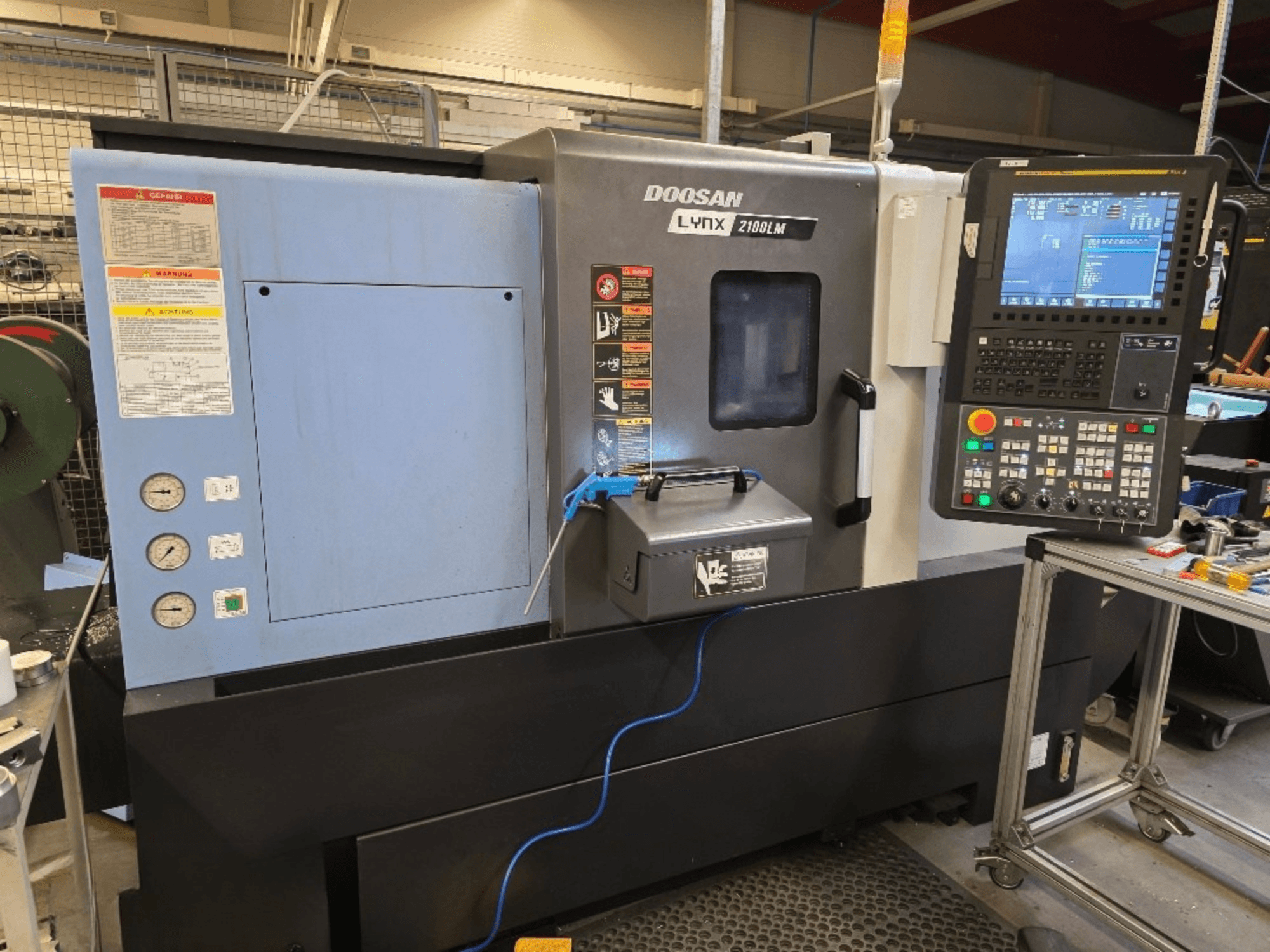 Токарный станок с ЧПУ Doosan Lynx 2100LM, вид спереди, панель управления, манометры и предупреждения о безопасности.