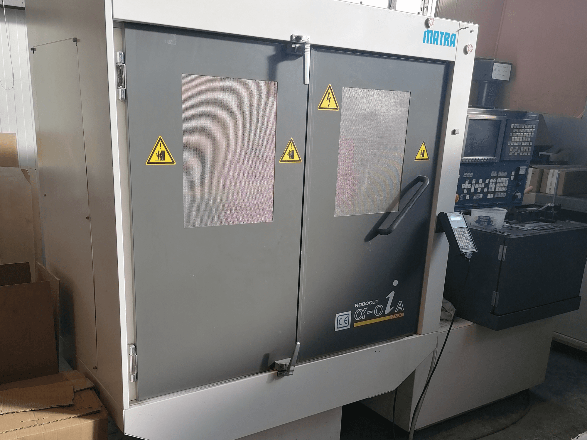 Станок Robocut α-oZα от Matra, вид спереди, где показаны предупреждения о безопасности, панель управления и двойные двери с сетчатыми окнами.