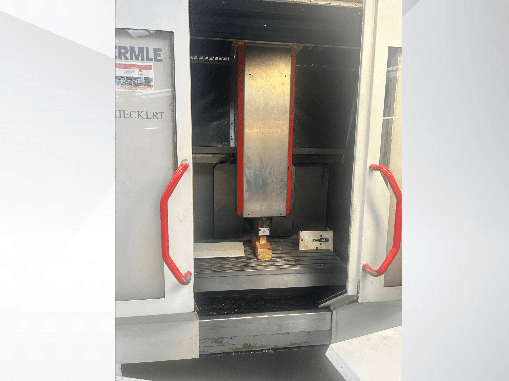 Вид спереди станка с ЧПУ Hermle с металлическим интерьером, красными рукоятками и заготовкой на столе.