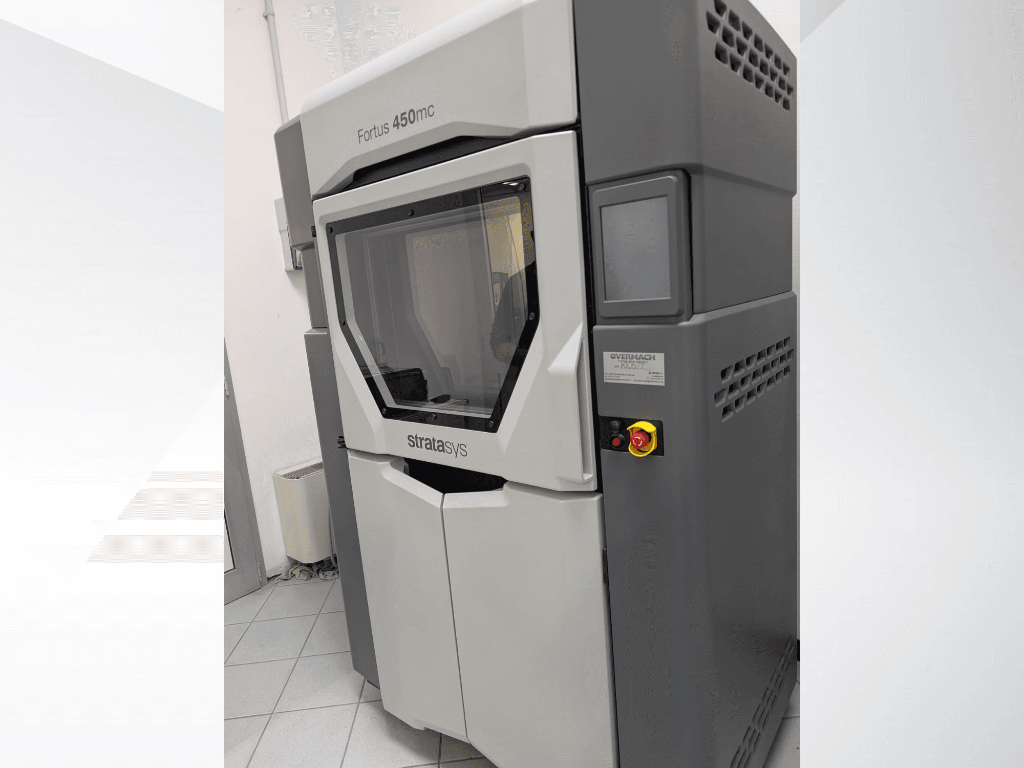 Пластиковый 3D-принтер Fortus 450mc показан сбоку, имеет прозрачную переднюю панель и кнопки управления сбоку.