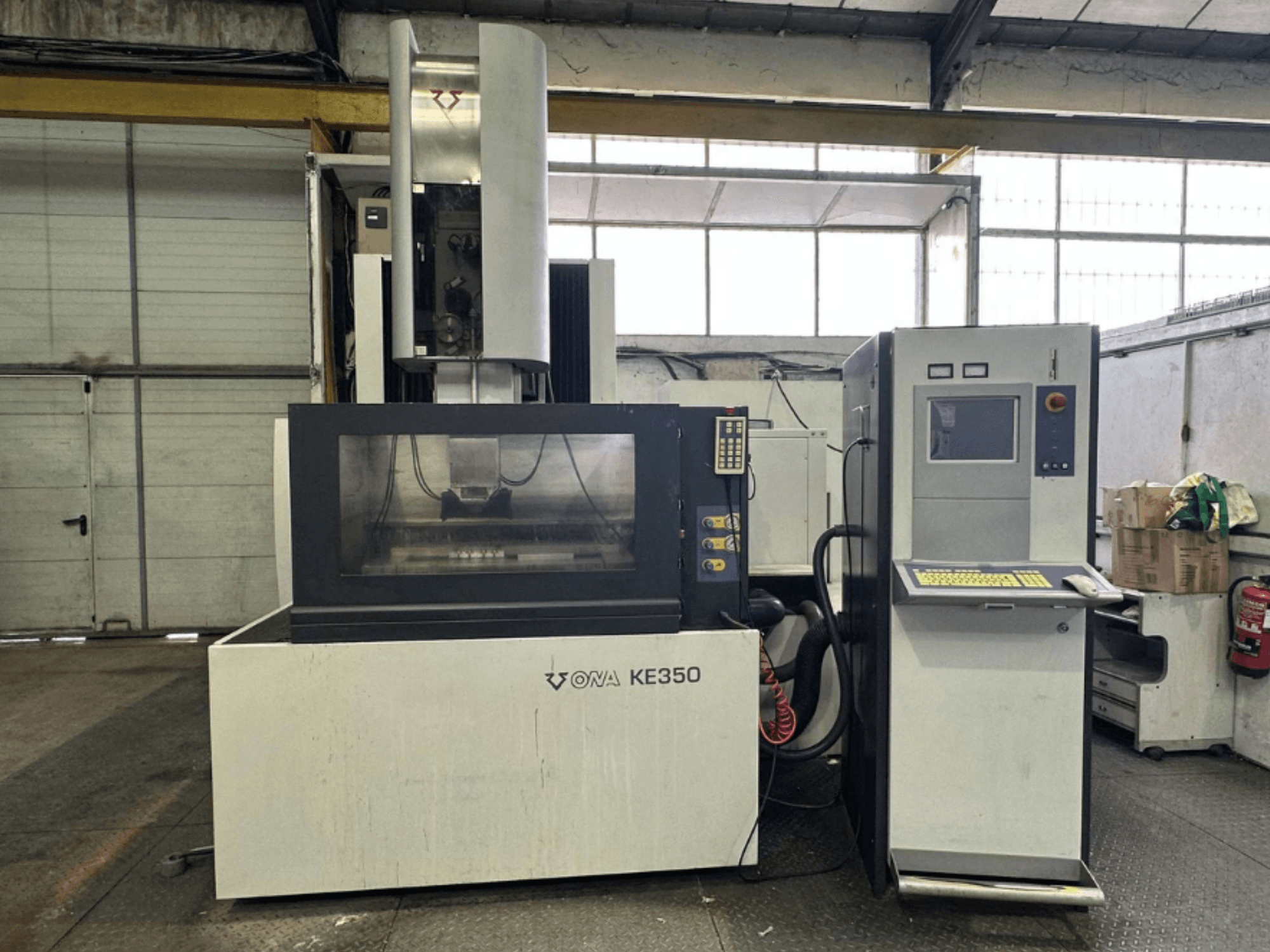 Вид спереди на станок с ЧПУ ONA KE350 с панелью управления, экраном и различными кнопками, расположенный в промышленном помещении.