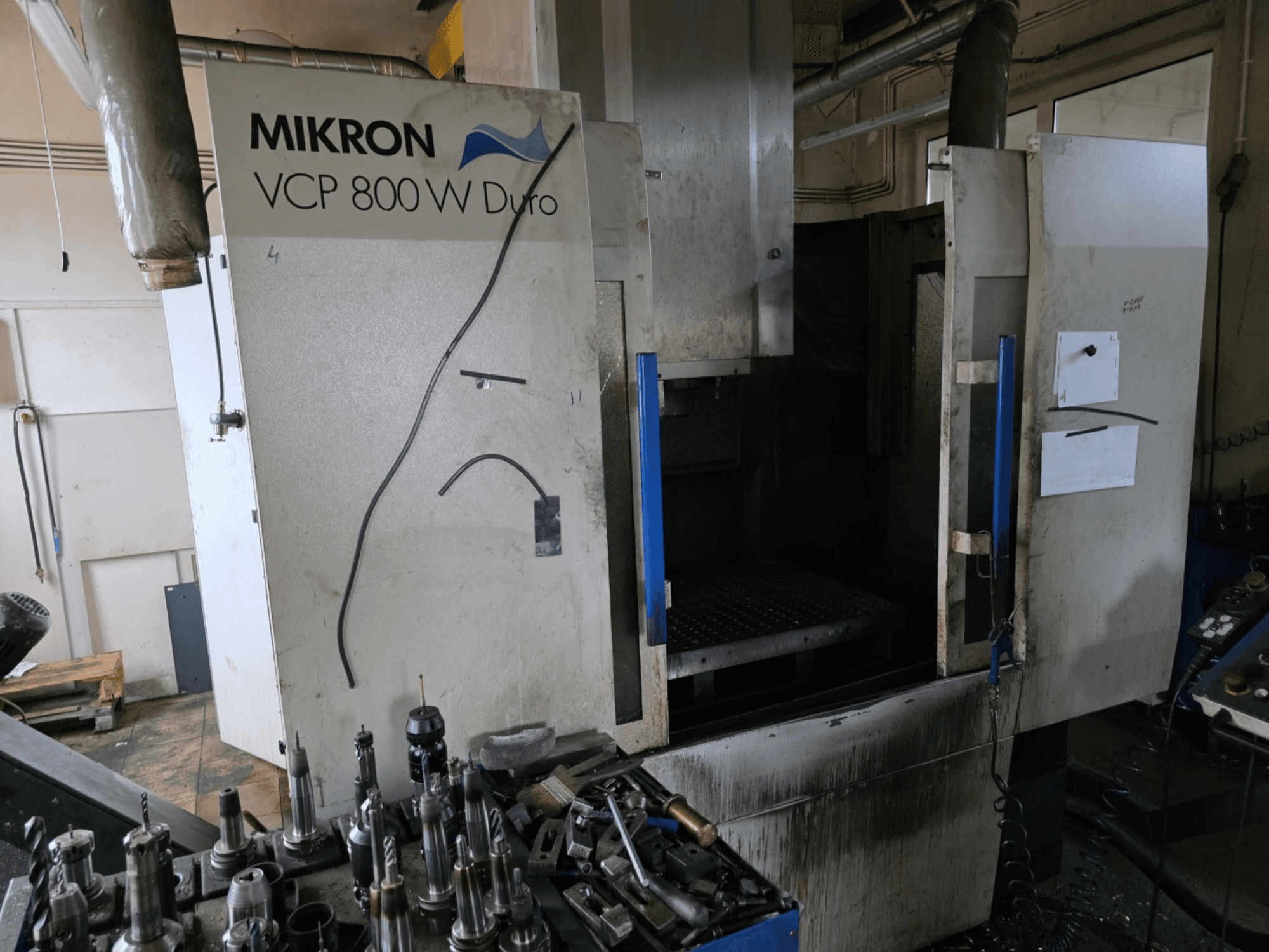 Фрезерный станок Mikron VCP 800 W Duro показан с боковой стороны, на переднем плане видны инструменты и органы управления.