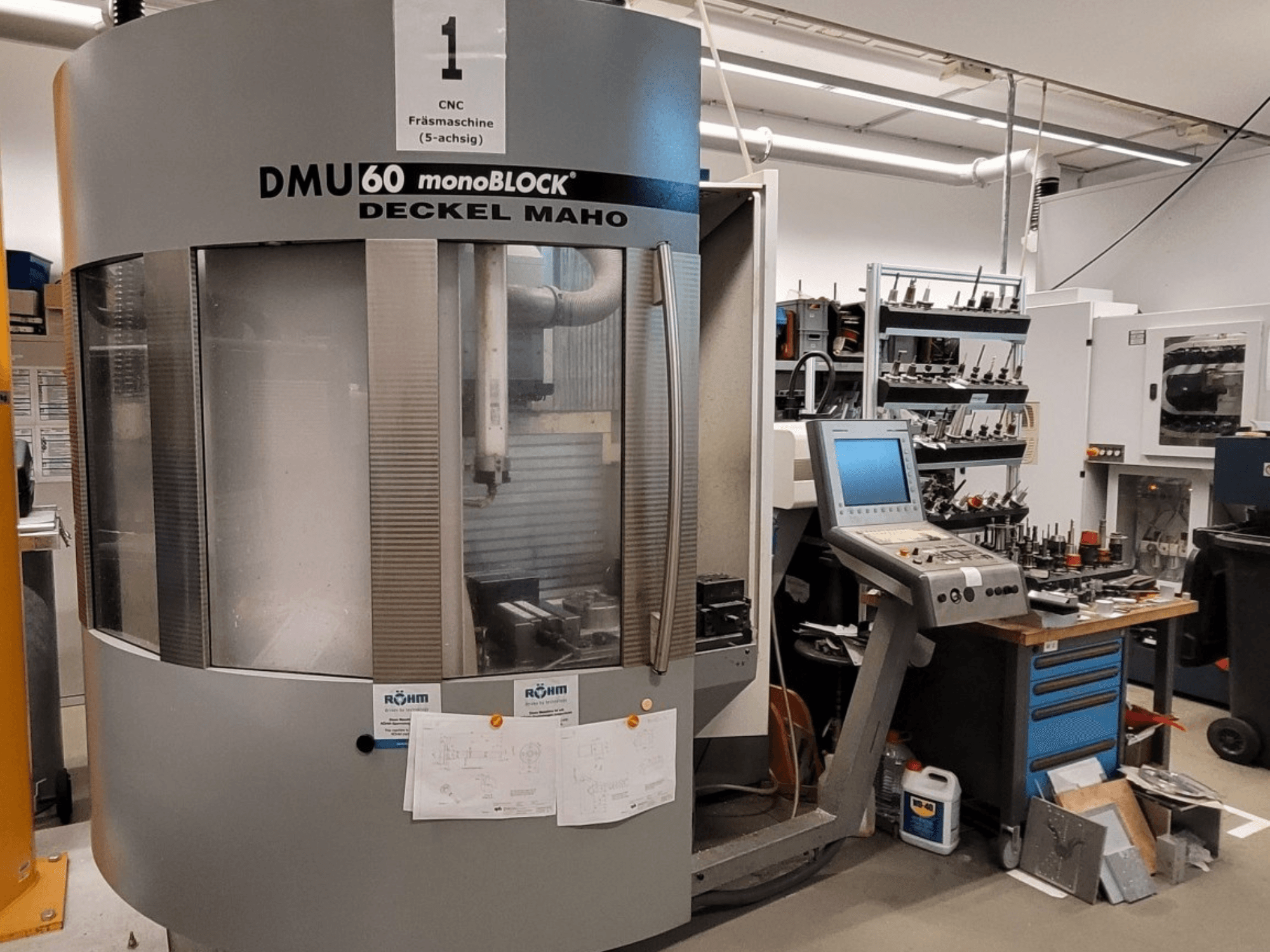 Фрезерный станок с ЧПУ DECKEL MAHO DMU 60 monoBLOCK на виде спереди, демонстрирует панель управления и хранилище инструментов рядом с ней.