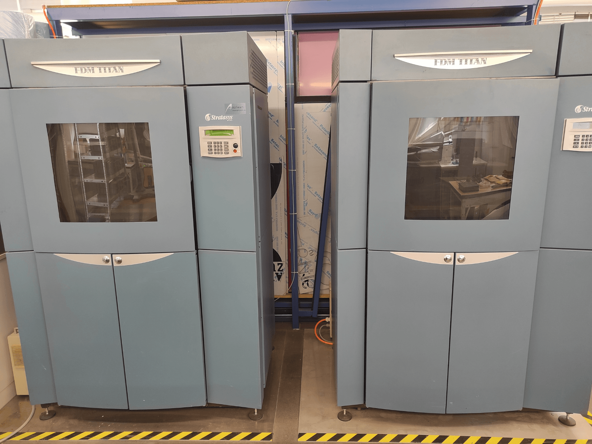 3D-принтер FDM TitanTI от Stratasys, вид спереди, с двумя дверцами и панелью управления, установленный в промышленном помещении.