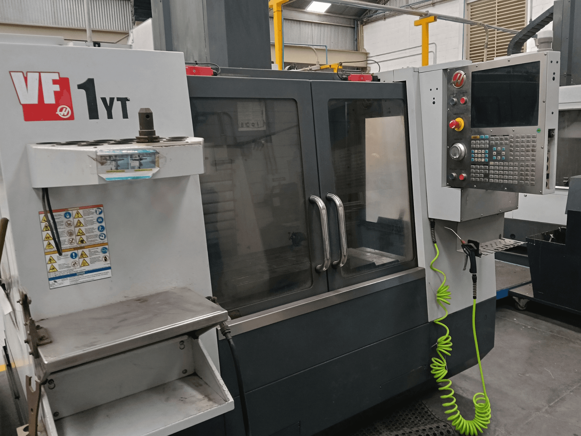 Вертикальный обрабатывающий центр HAAS VF-1YT-2, вид спереди, на котором видны панель управления, держатель инструмента, предупреждающие надписи и зона обработки.