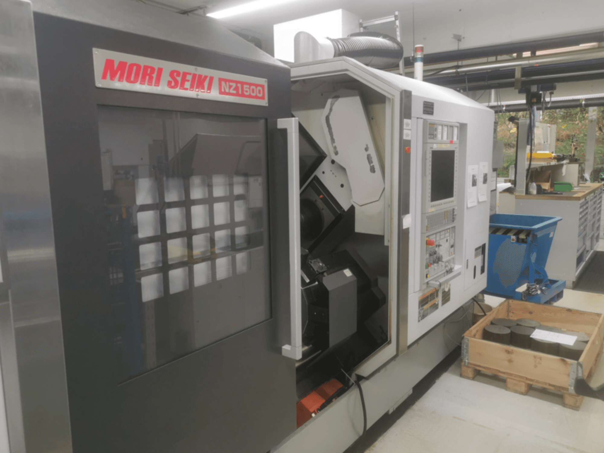 Станок с ЧПУ MORI SEIKI NZ1500 под небольшим углом, видна панель управления и рабочая зона с металлическими заготовками на деревянном поддоне.