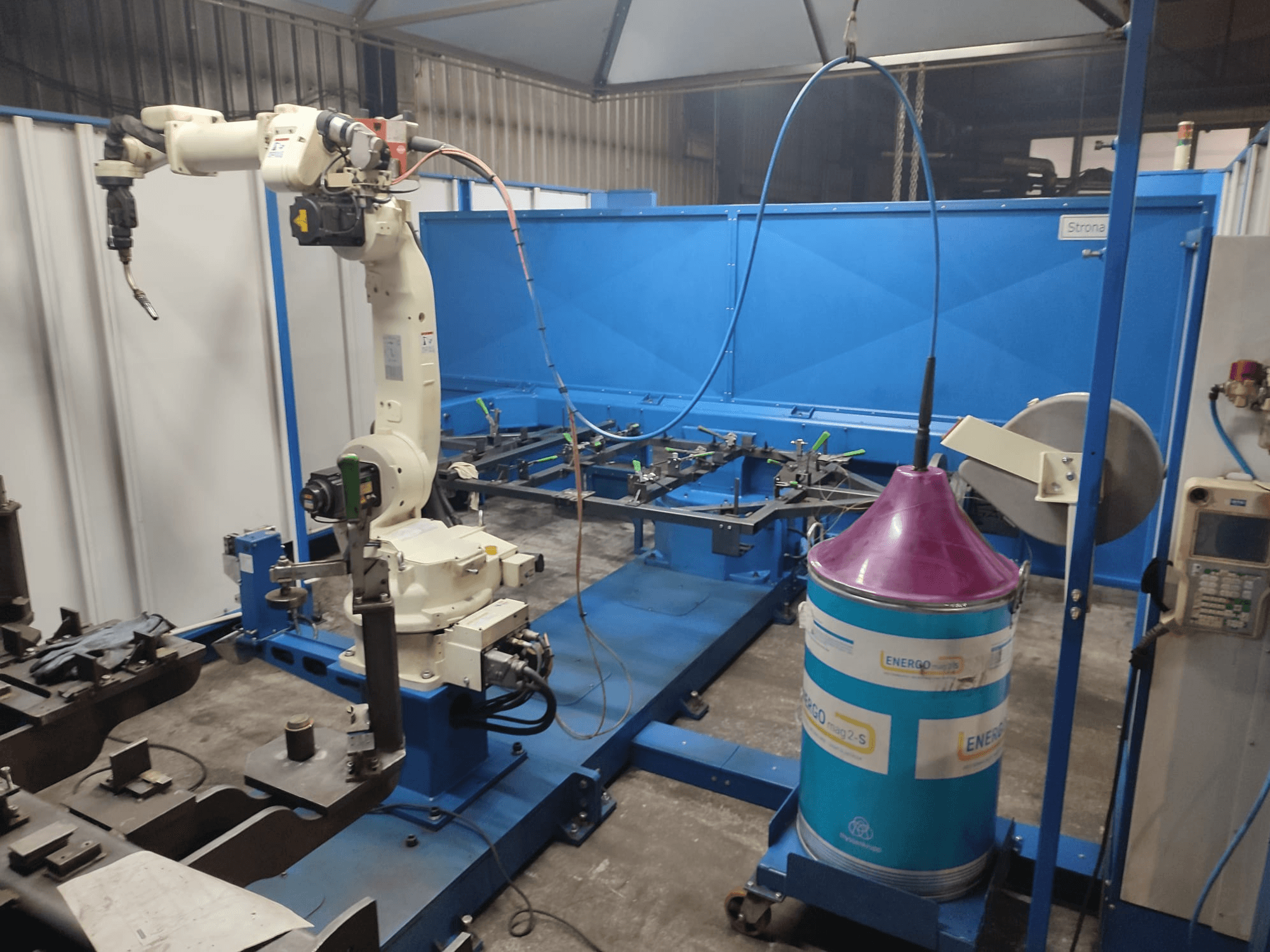 Промышленная роботизированная рука с бело-голубым основанием, установленная в мастерской с синей рабочей поверхностью и металлическими компонентами.