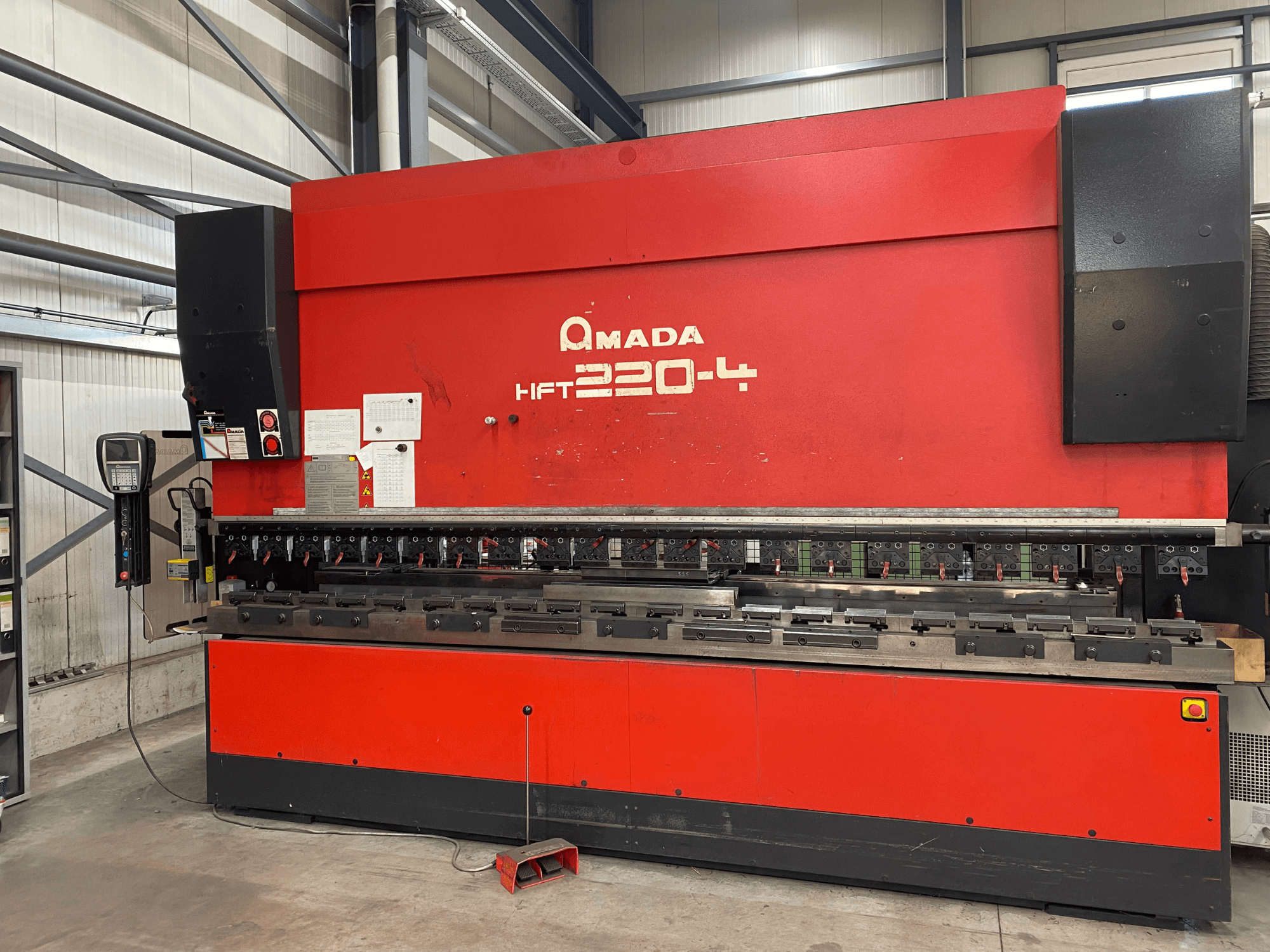 Красный листогибочный пресс AMADA HFT 220-4 в заводских условиях, вид спереди на панель управления и гибочные инструменты на рабочей поверхности.
