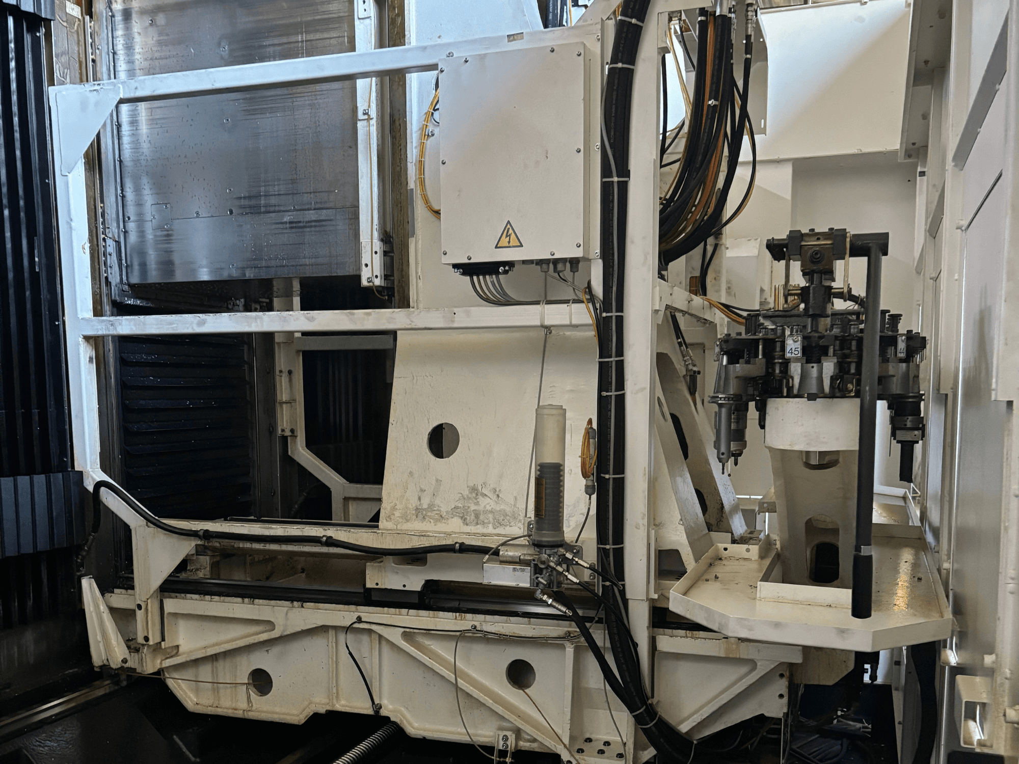 Вид сбоку на промышленный станок, на котором изображен белый фрезерный станок с ЧПУ с видимыми инструментами, трубами и панелью управления.