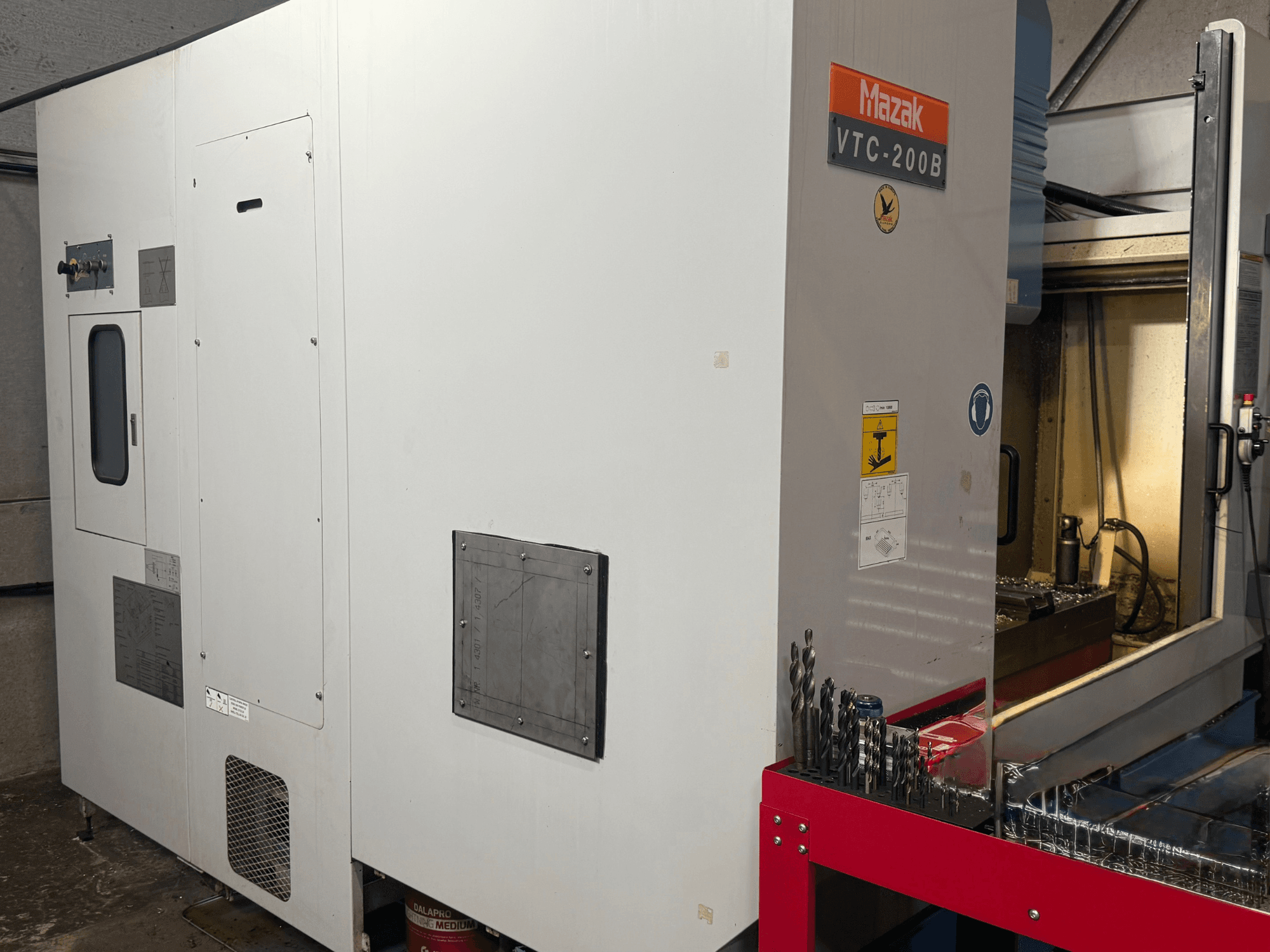 Вертикальный обрабатывающий центр Mazak VTC-200B в боковом виде с обозначенными элементами управления и сверлами на красной стеллажной полке.