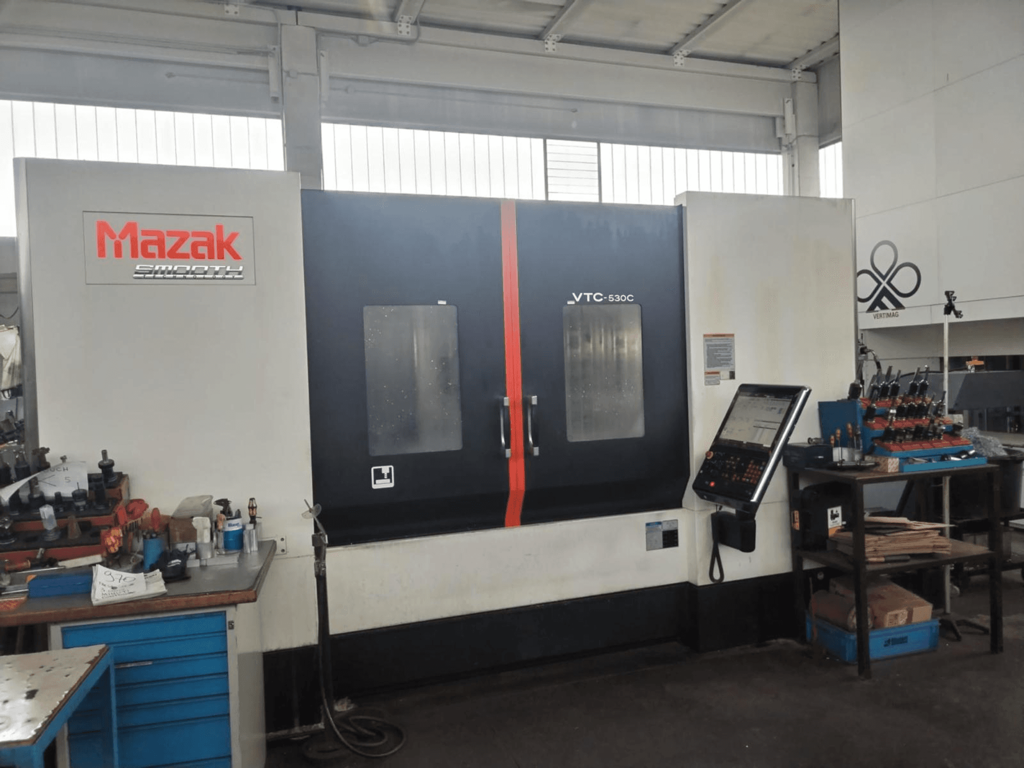 Вертикальный обрабатывающий центр Mazak VTC-530C в цехе с панелью управления и инструментами на боковом столе.
