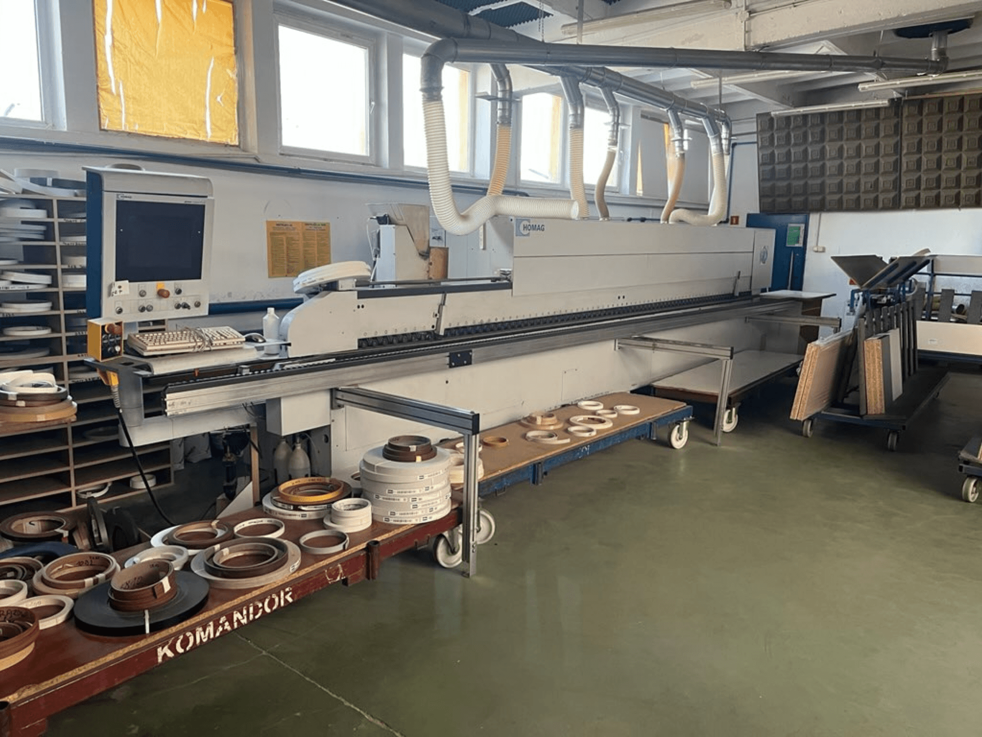 Кромкооблицовочный станок HOMAG в мастерской, элементы управления и различные материалы для кромкооблицовки на синей тележке. Вид сбоку.