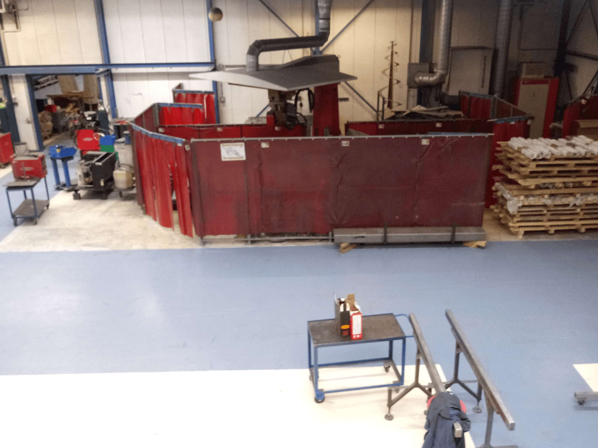 Промышленное рабочее место с красными сварочными шторами, станками и поддонами; вид сверху, демонстрирующий расположение и оборудование.