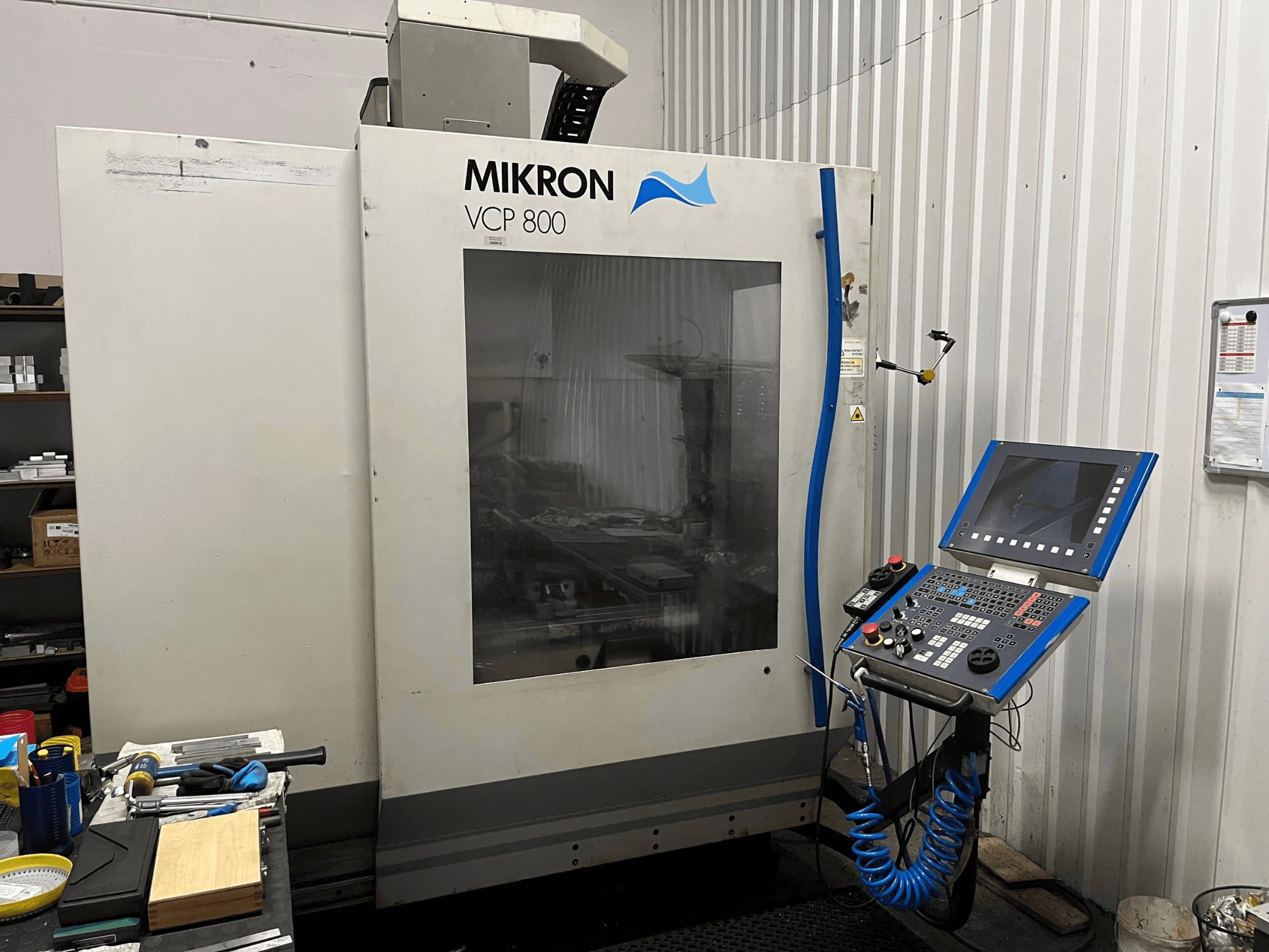 Вертикальный обрабатывающий центр MIKRON VCP 800, вид спереди, с панелью управления и инструментами на рабочем столе на переднем плане.