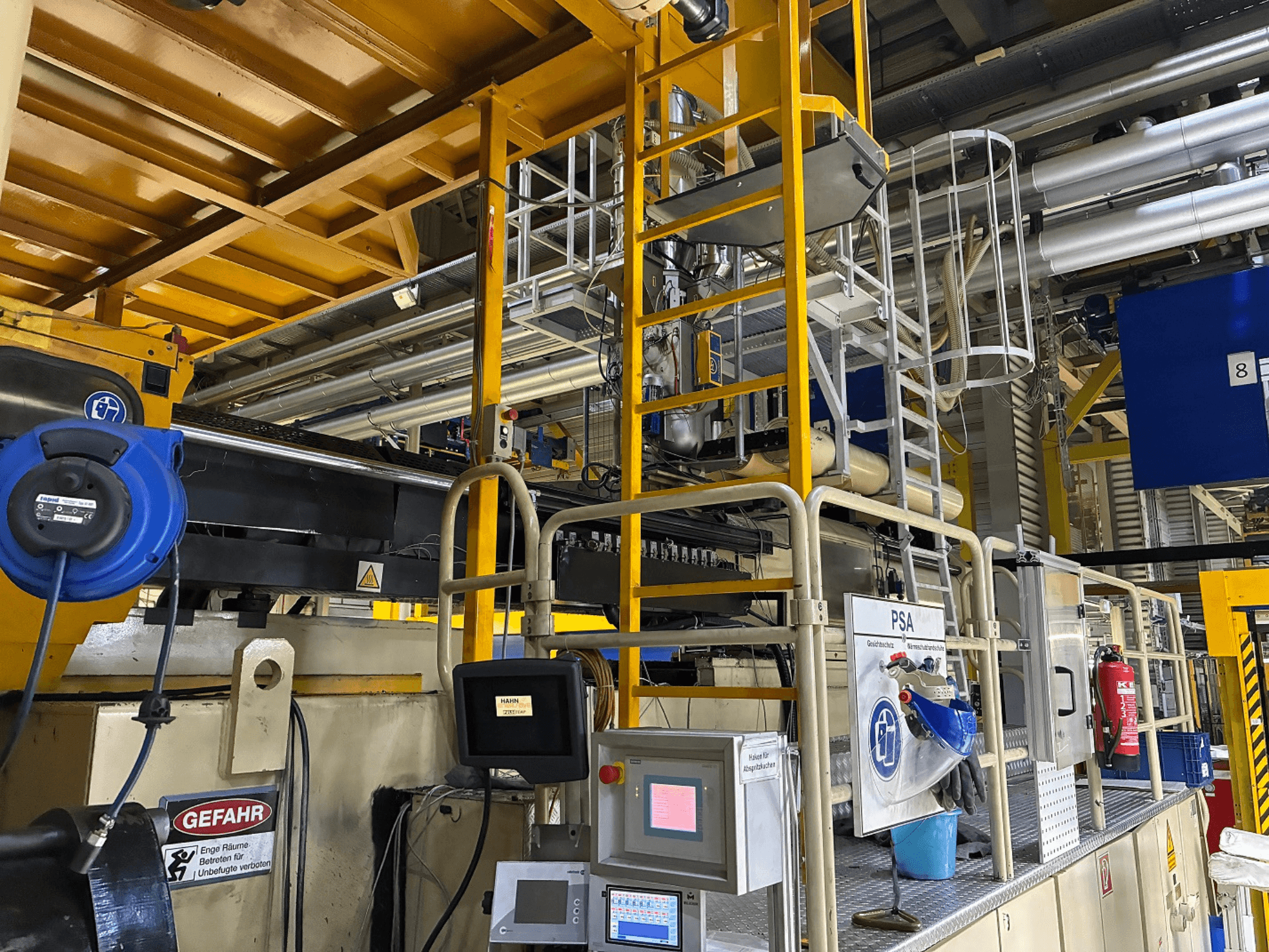 Пульт управления промышленным оборудованием с синими средствами безопасности и желтыми строительными лесами, вид сбоку с изображением труб и предупреждающих знаков.