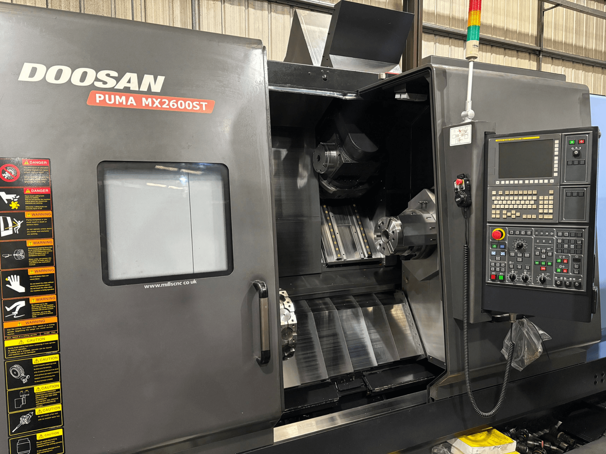 Токарный станок с ЧПУ Doosan PUMA MX2600ST, вид спереди, показывает панель управления, шпиндель и зону обработки с видимыми предупреждениями о безопасности.