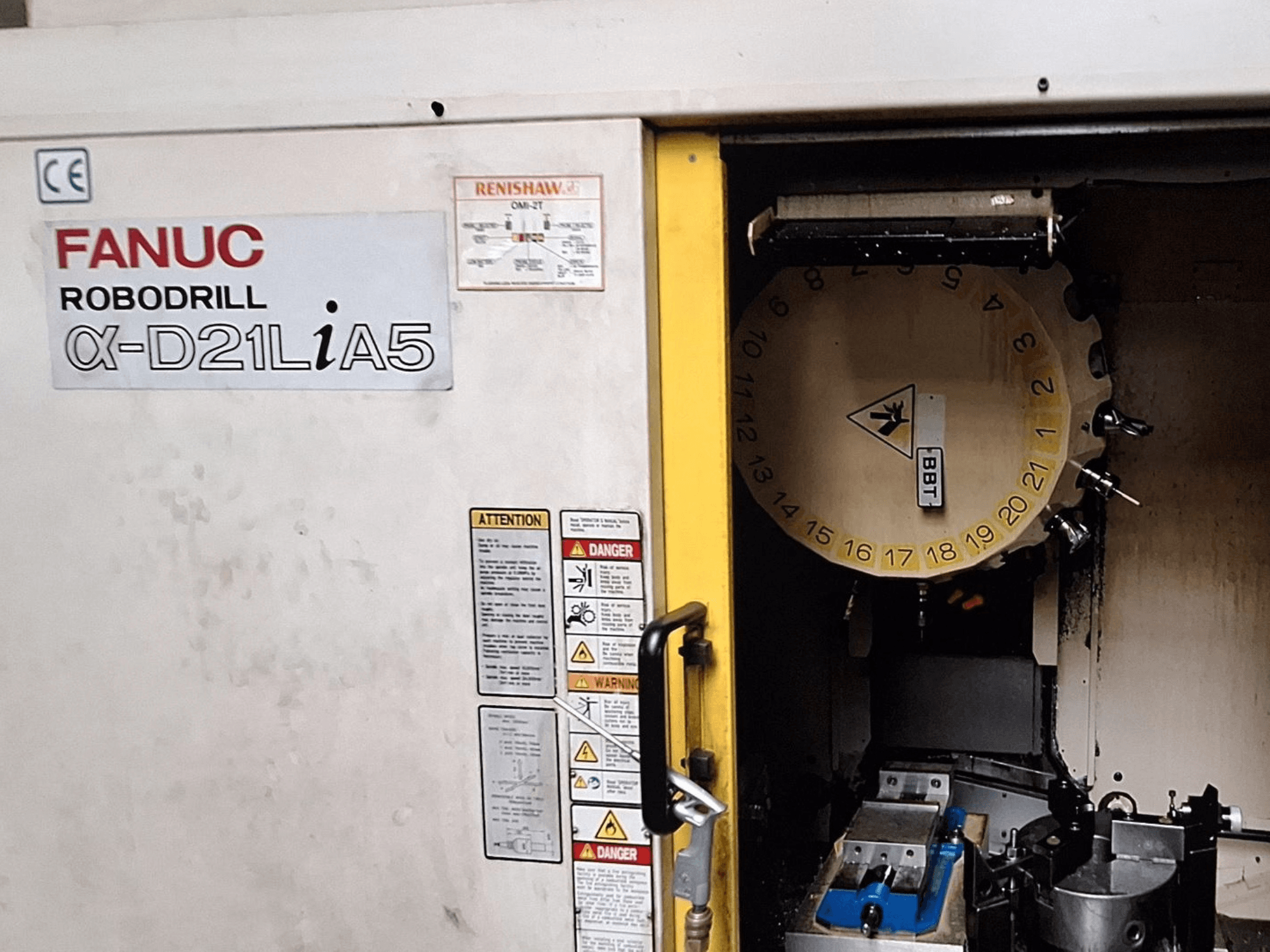 FANUC Robodrill α-D21LiA5, вид спереди. Показывает карусель с инструментами и зону обработки с зажимами и предупреждениями на дверце.