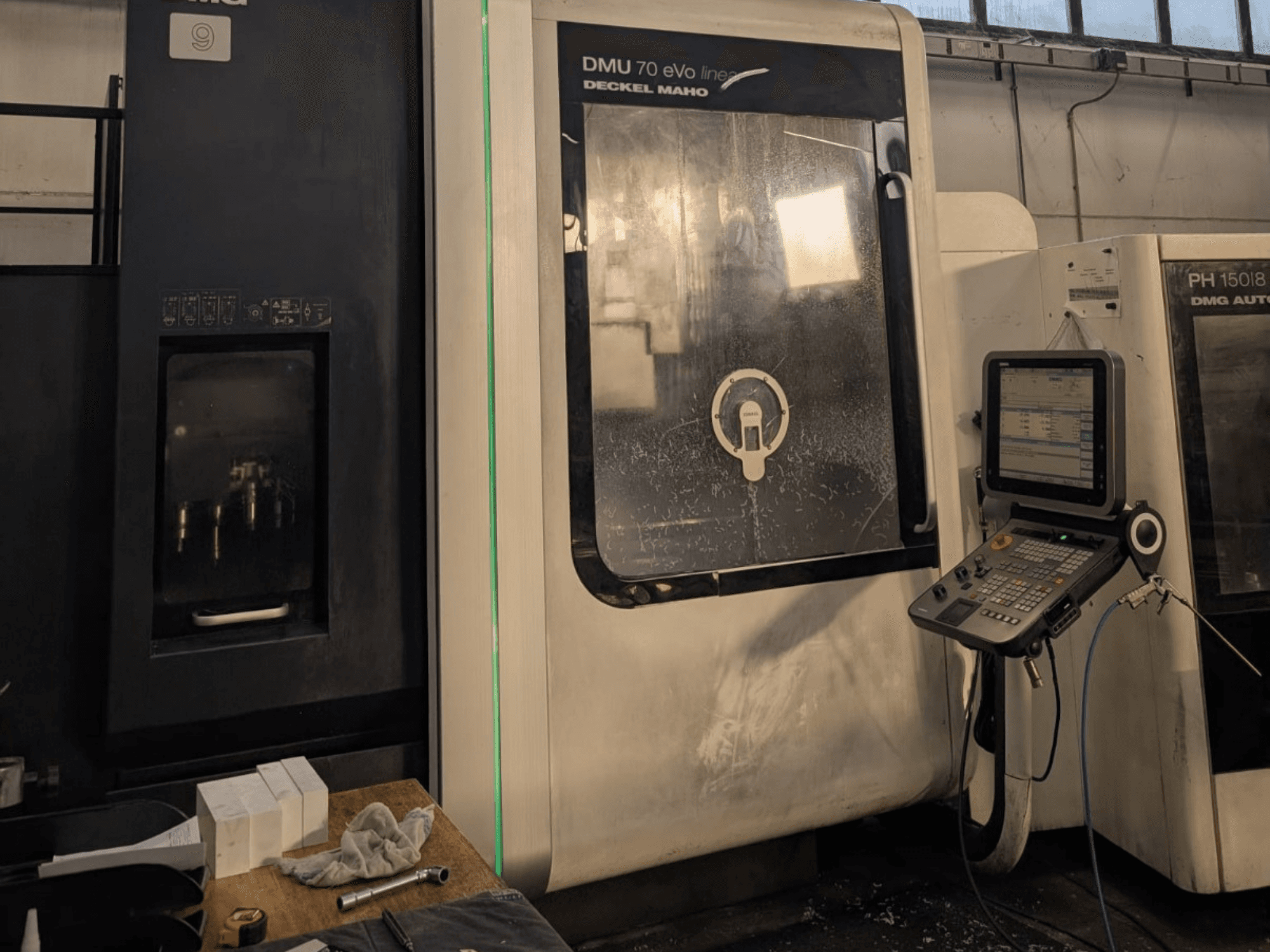 Линейный вертикальный обрабатывающий центр DMG DECKEL MAHO DMU 70 EVO, вид спереди с панелью управления, инструментами и рабочей зоной.
