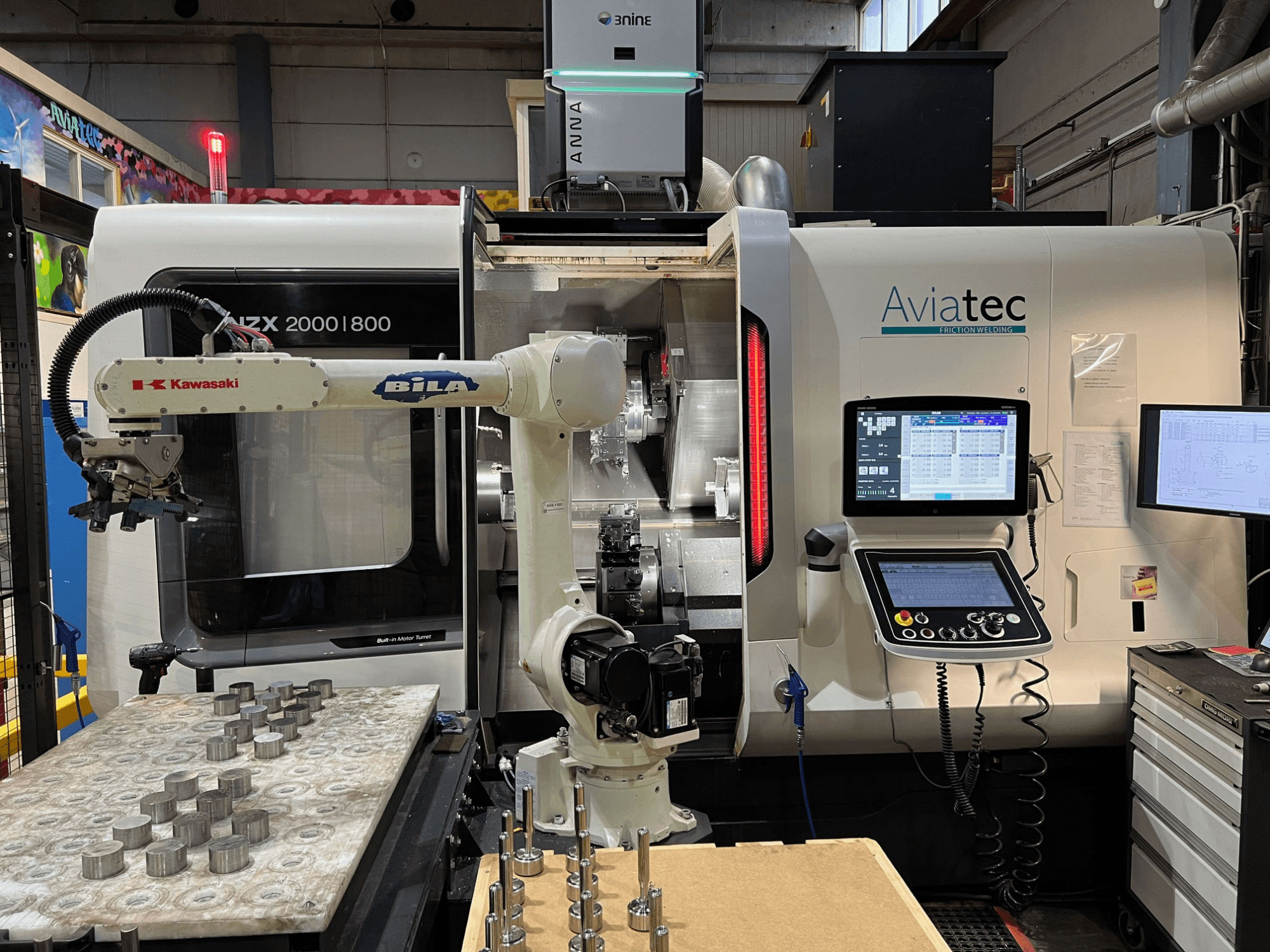 Горизонтальный токарный станок DMG MORI NZX 2000/800SY с роботом-манипулятором Kawasaki, пультом управления и металлическими деталями на рабочем столе.