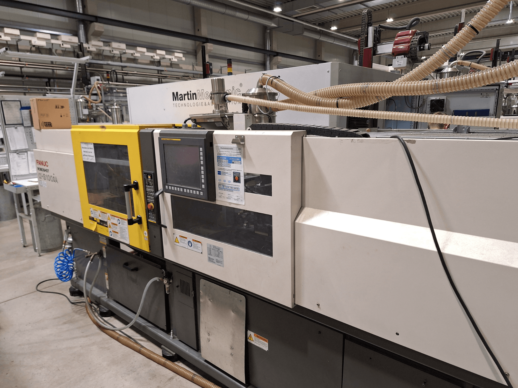 Электрическая машина для литья под давлением FANUC Roboshot S100iA, вид сбоку, оснащена сенсорным экраном и наклейками безопасности.