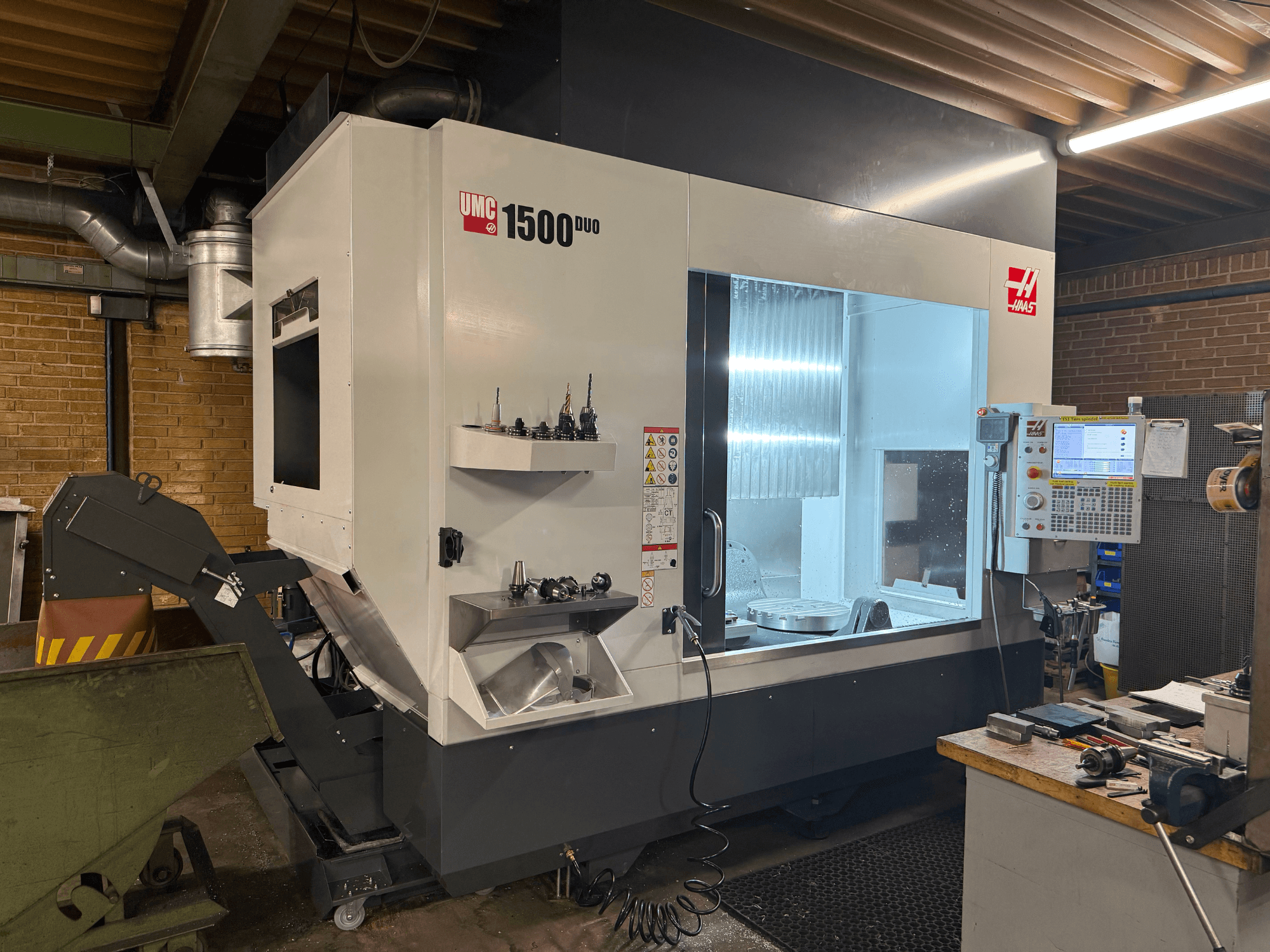 Станок с ЧПУ Haas UMC 1500DUO, вид спереди, имеет большое окно, панель управления и держатели инструментов на полке.