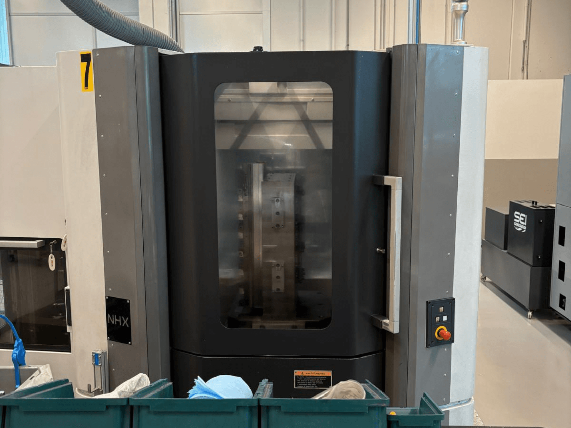 Станок с ЧПУ NHX (вид спереди), панель управления и наглядный дисплей; расположен в цеху, рядом зеленые контейнеры.