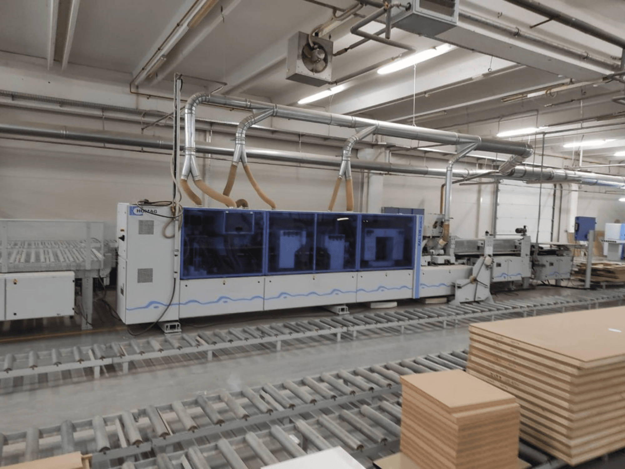 Производственный станок HOMAG на заводе, вид спереди, с синим корпусом и конвейерной системой со штабелями фанерных листов.