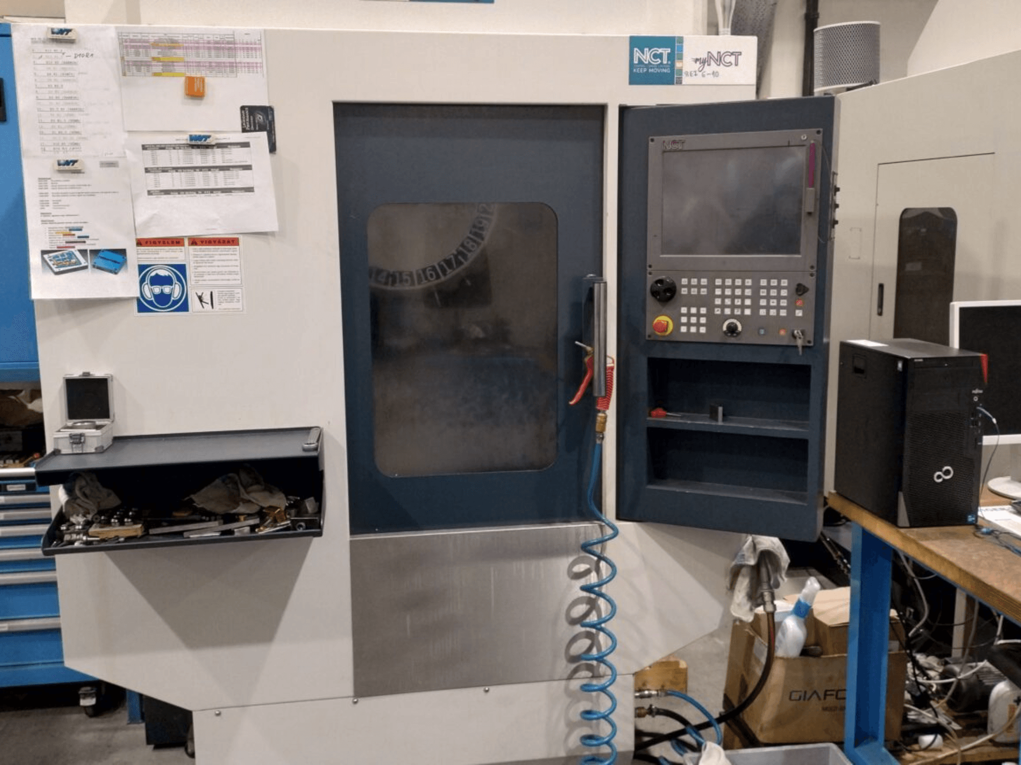 Станок с ЧПУ NCT, вид спереди, панель управления с цифровой клавиатурой, инструменты в ящике для хранения, руководство на стене.