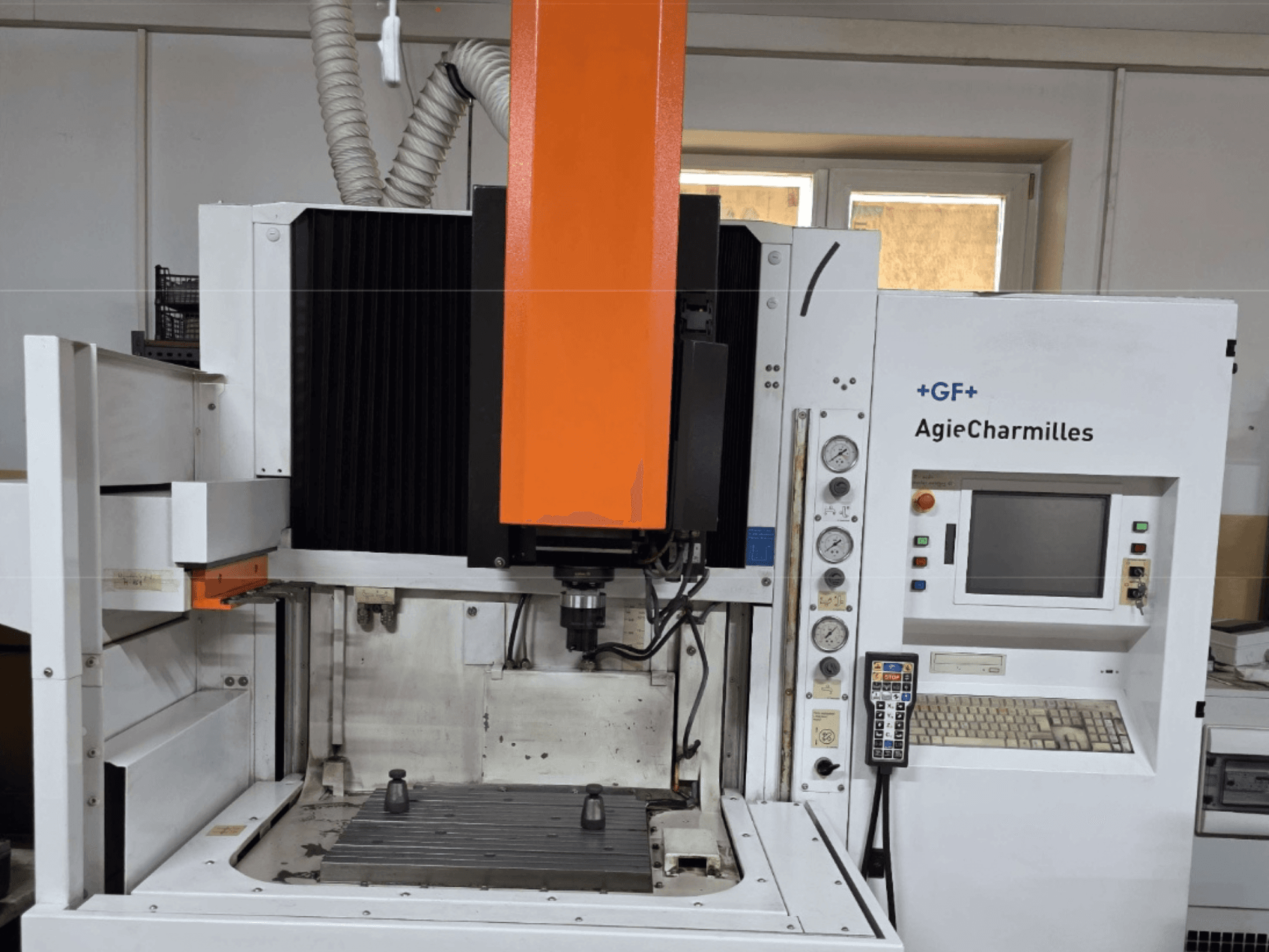 Обрабатывающий центр AgieCharmilles на переднем плане с большим оранжевым шпинделем, панелью управления и видимым рабочим столом.