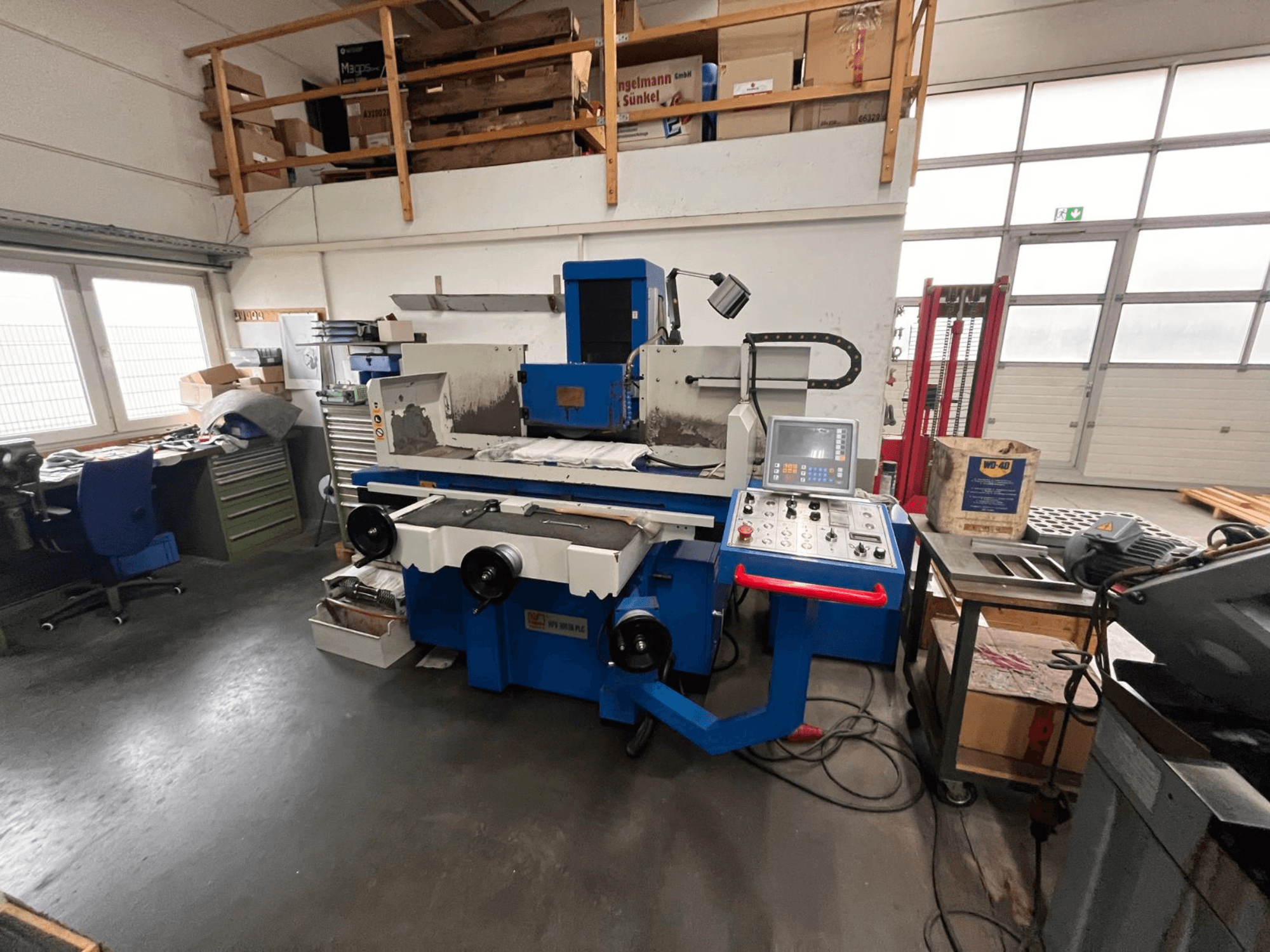 Синий плоскошлифовальный станок с цифровой панелью управления, вид спереди, на фоне загромождения рабочего пространства и зеленого шкафа для инструментов.