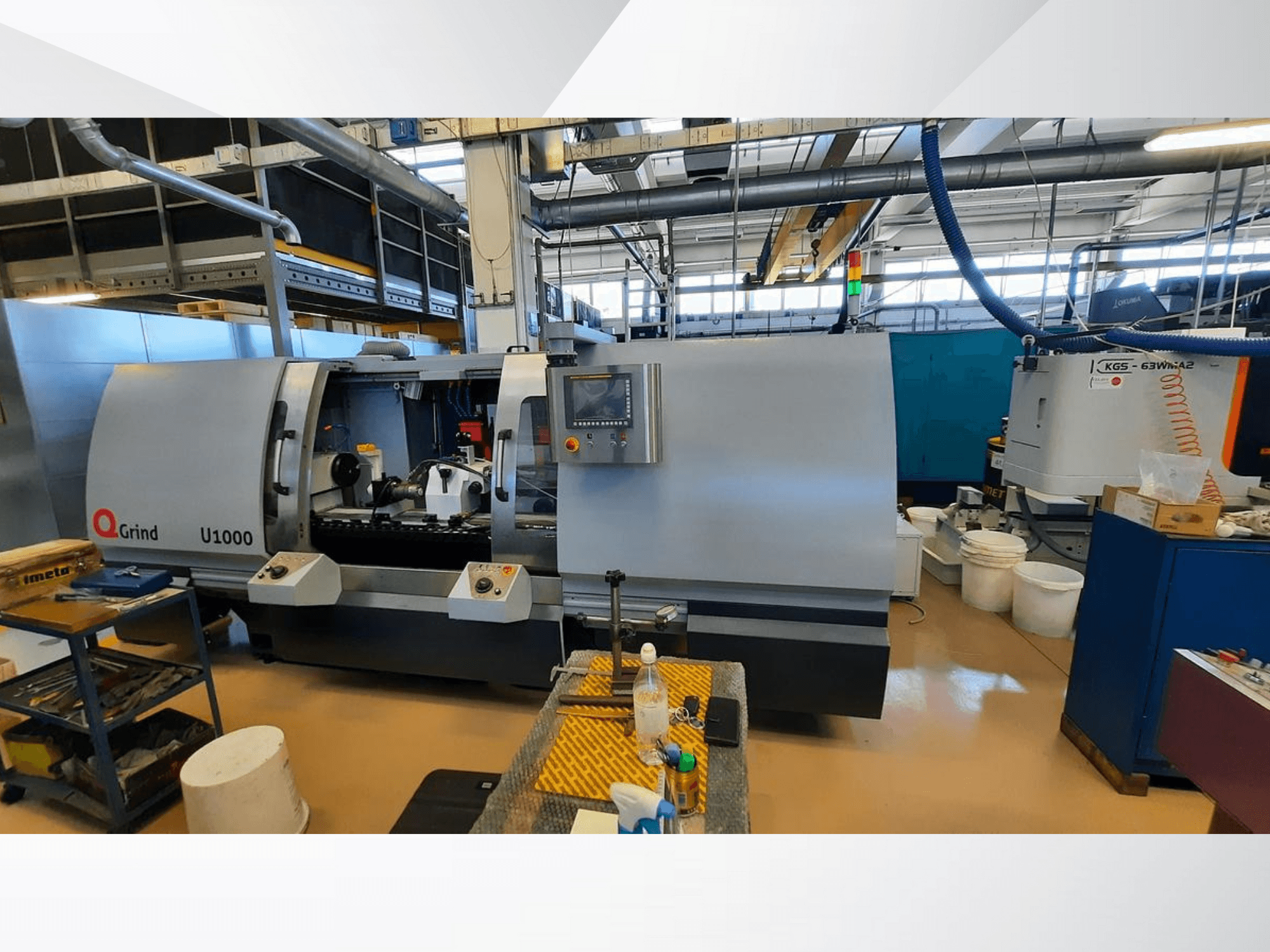 Станок Q-Grind U1000 в мастерской, вид спереди с панелью управления и открытой зоной резки, окруженной оборудованием.