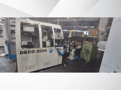 Станок DECO 2000 спереди: панель управления, трубы и загроможденное инструментами и коробками рабочее место.