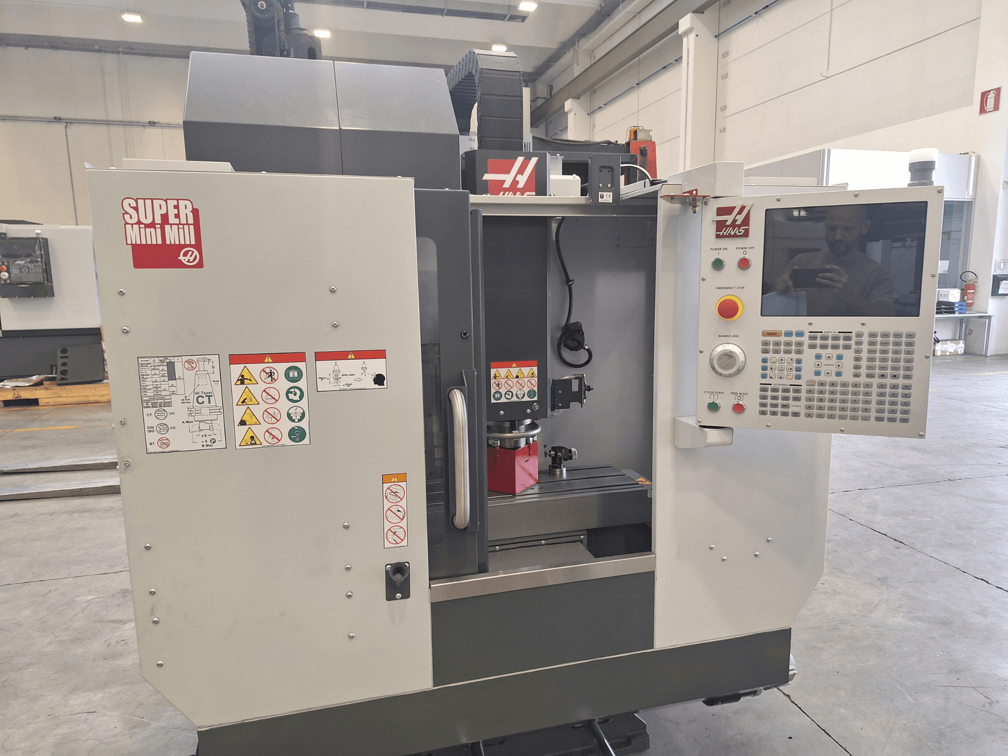 Вид спереди фрезерного станка HAAS Super Mini Mill с панелью управления, предупреждающими надписями и зоной обработки с установленной заготовкой.