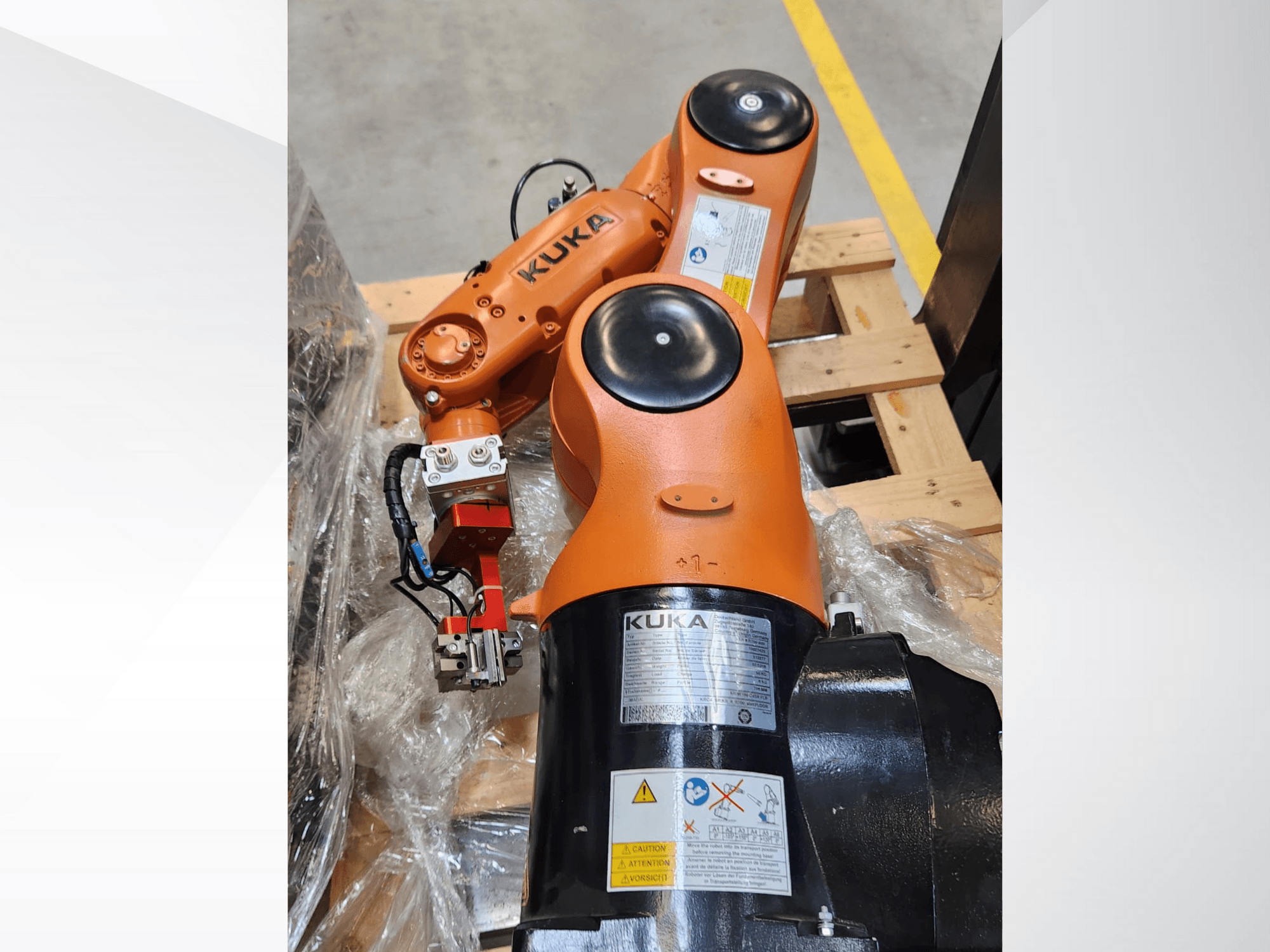 Роботизированная рука KUKA с оранжевой отделкой, вид сбоку, с захватом и проводами управления на деревянном поддоне.