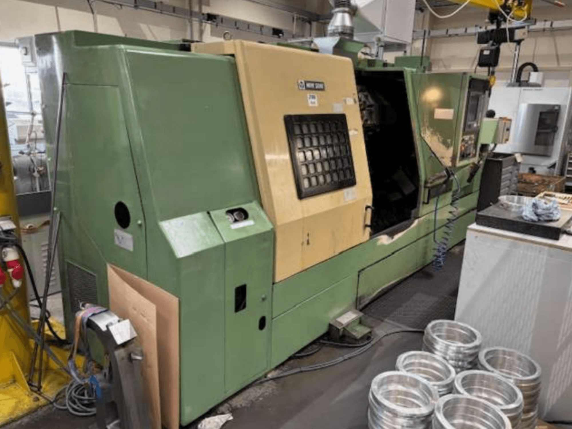 Зеленый токарный станок с ЧПУ Mazak, вид спереди, демонстрирует панель управления и обработанные алюминиевые детали на рабочем столе.