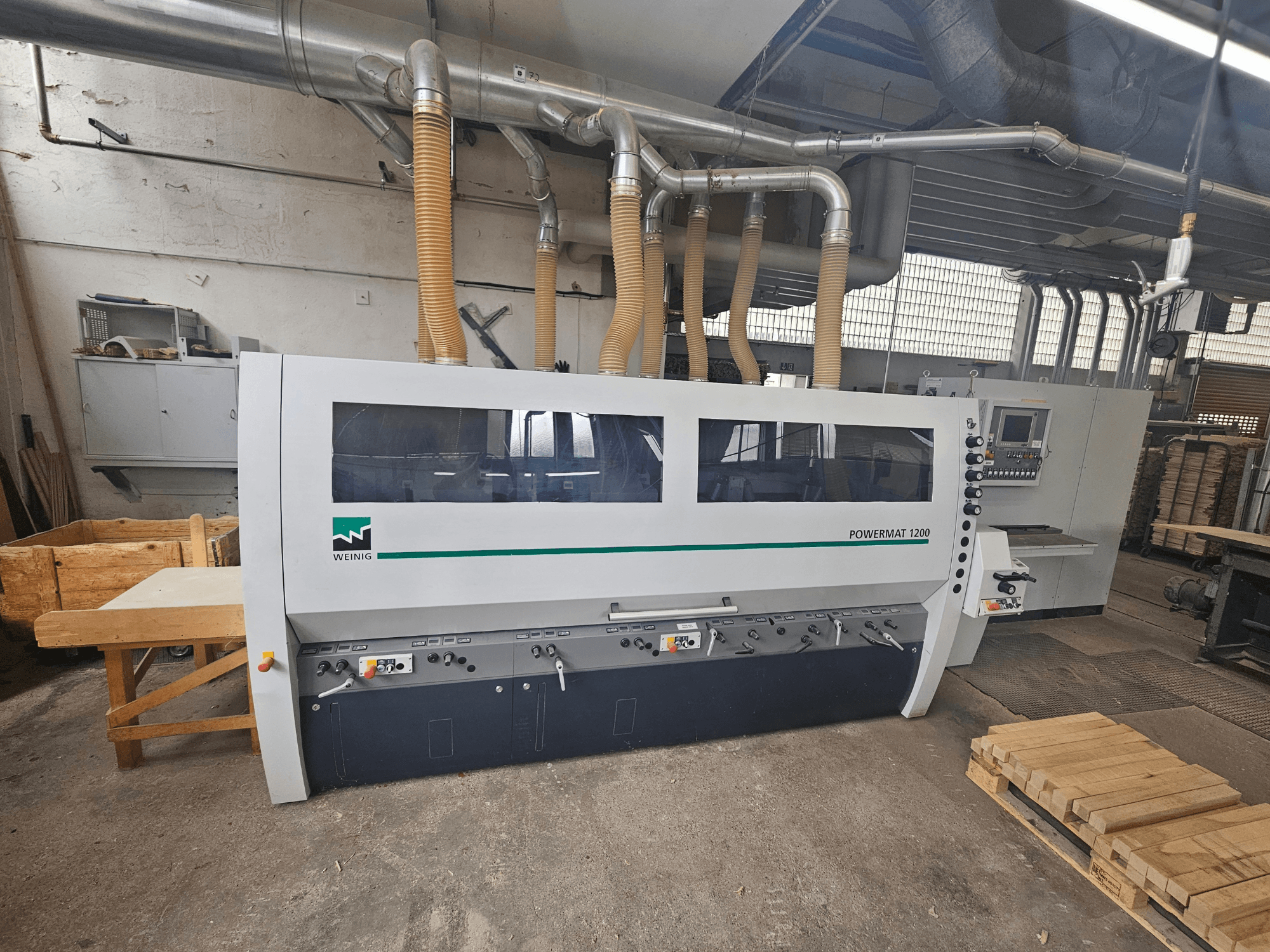 Деревообрабатывающий станок Weinig Powermat 1200, вид спереди, с панелью управления и штабелями древесины в мастерской.