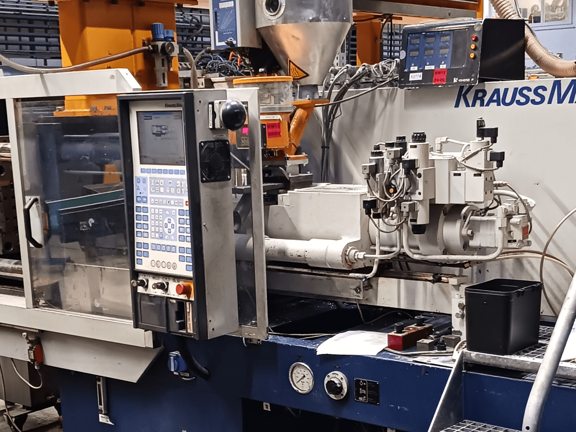 Термопластавтомат KraussMaffei с панелью управления, гидравлическими узлами и инструментами на переднем плане, вид спереди.