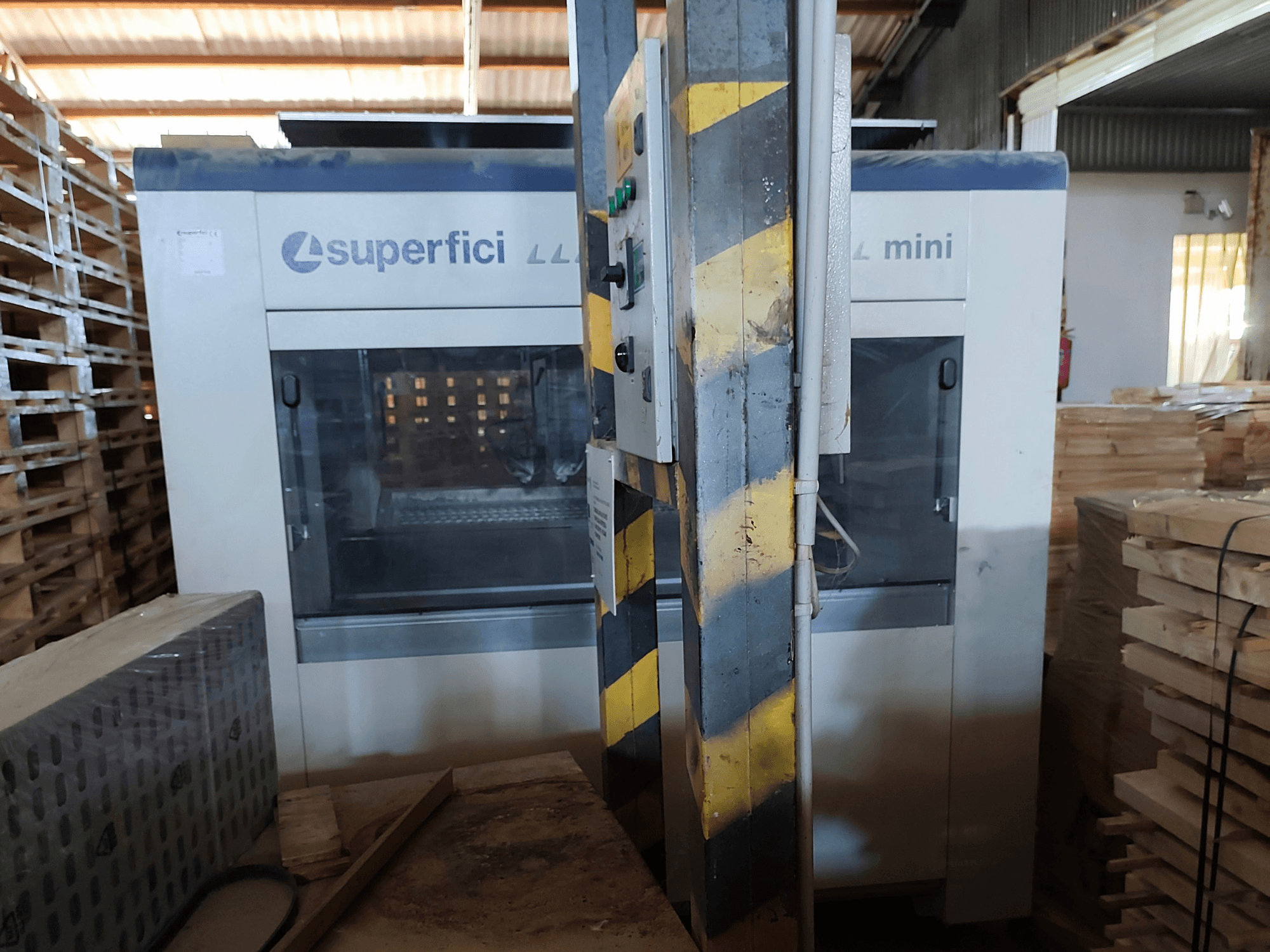 Деревообрабатывающий станок SCM Superfici Mini в виде спереди, на котором видны панель управления и окружающие деревянные поддоны.