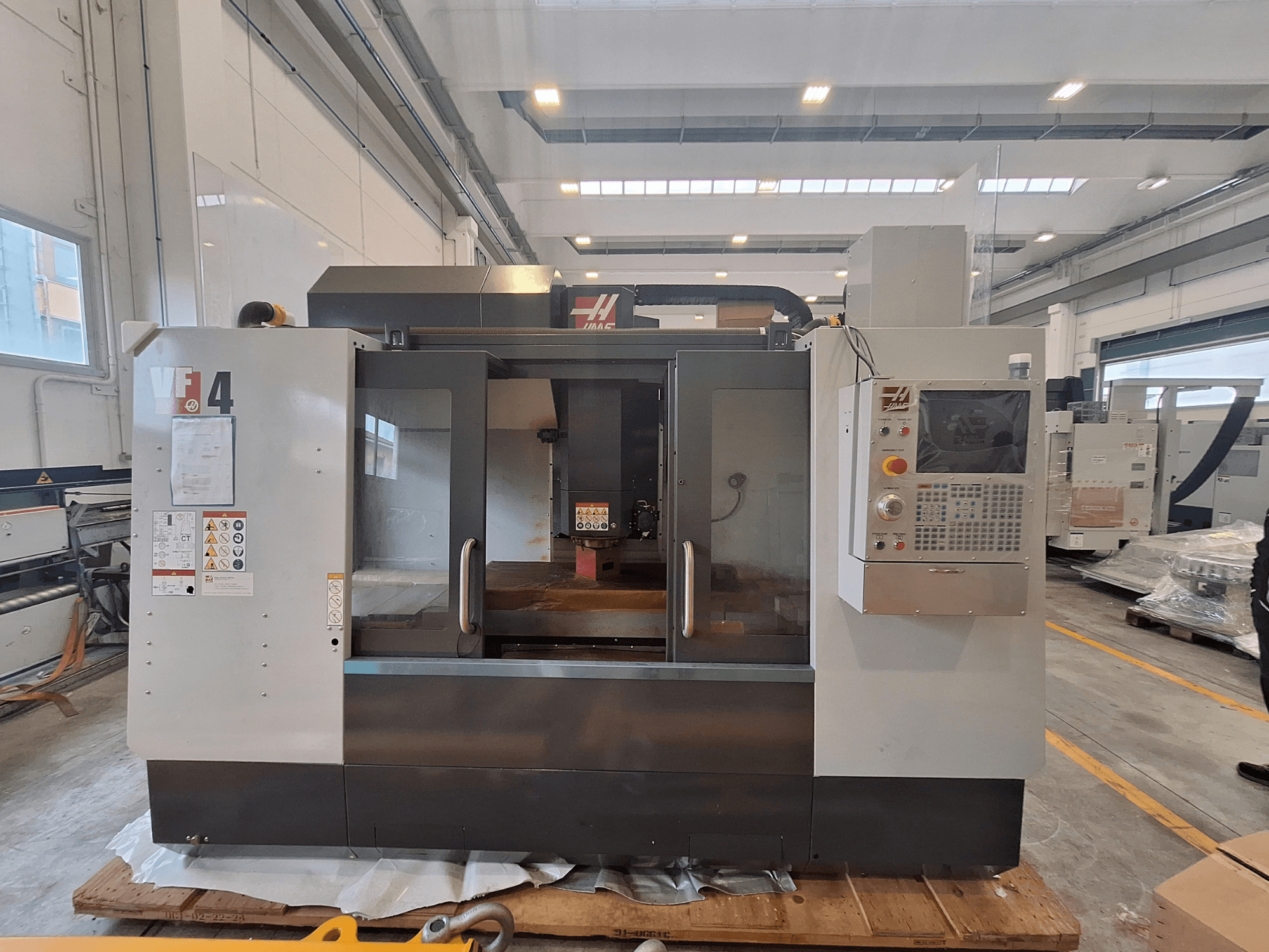 Вертикальный обрабатывающий центр HAAS VF-4, вид спереди, на котором видны панель управления, рабочая зона и предупреждающие надписи в условиях склада.