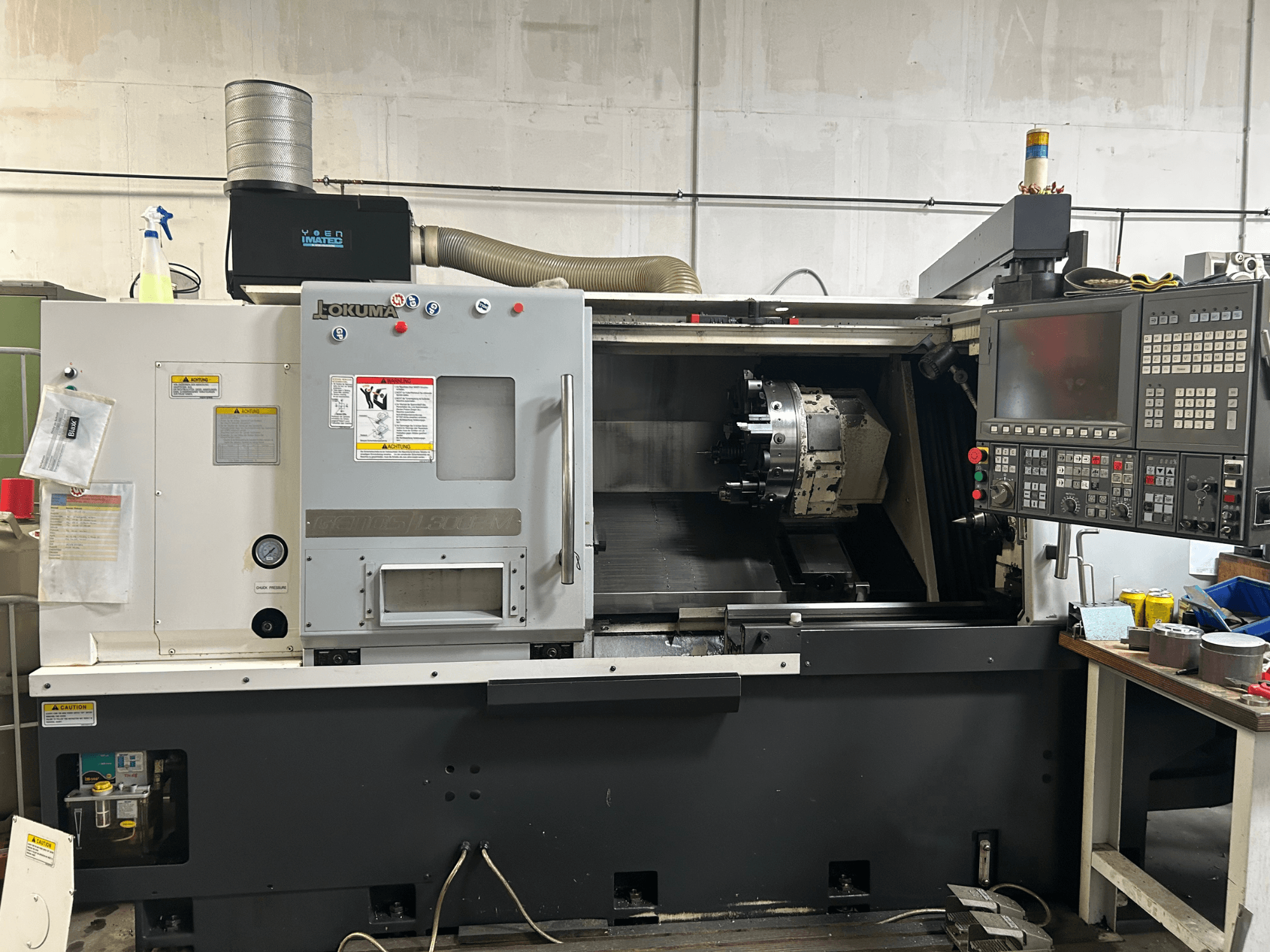 Вид спереди горизонтального токарного станка Okuma Genos L300E-M с панелью управления, видимым шпинделем и различными инструментами для настройки.