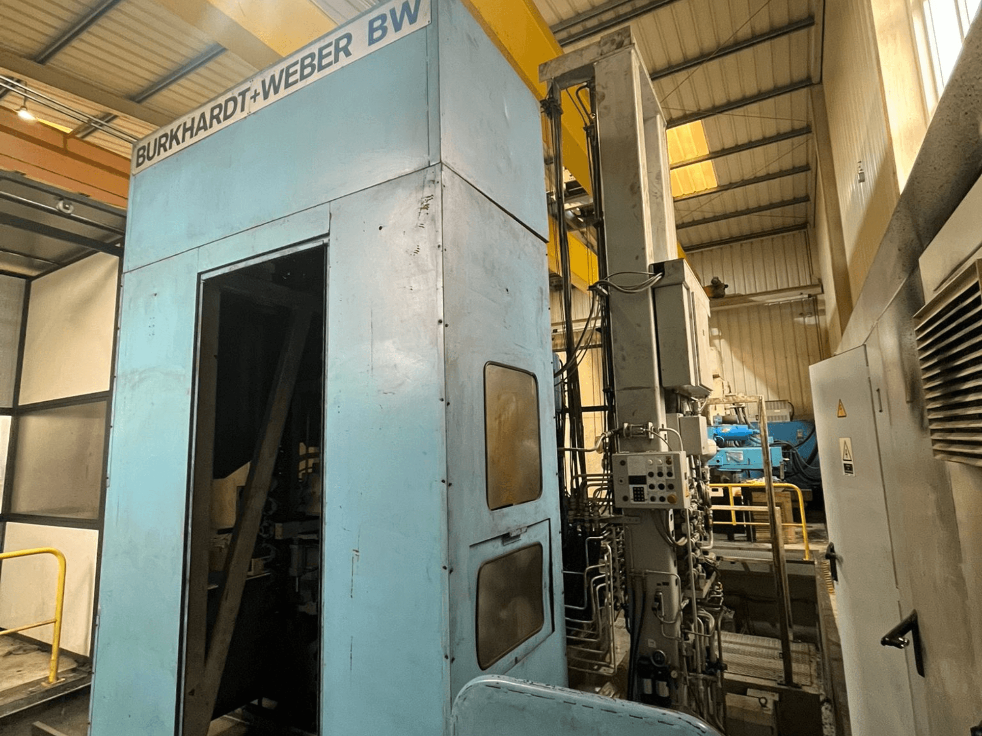 Горизонтальный обрабатывающий центр Burkhardt-Weber MC100, вид сбоку, на котором виден синий корпус и панель управления с проходящими трубопроводами.