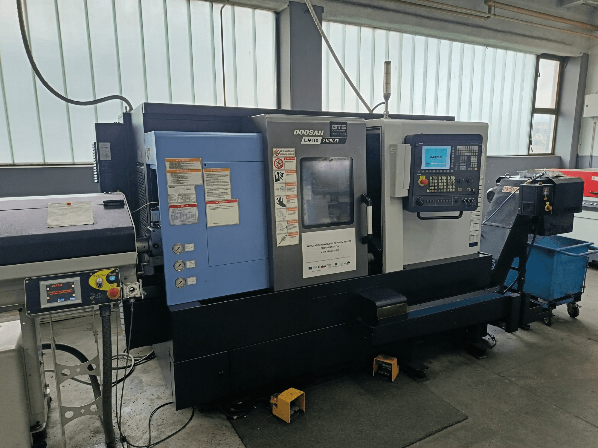 Токарно-фрезерный центр DOOSAN LYNX 2100 LSY, вид сбоку с изображением панели управления и кнопок управления, видны информационные наклейки.