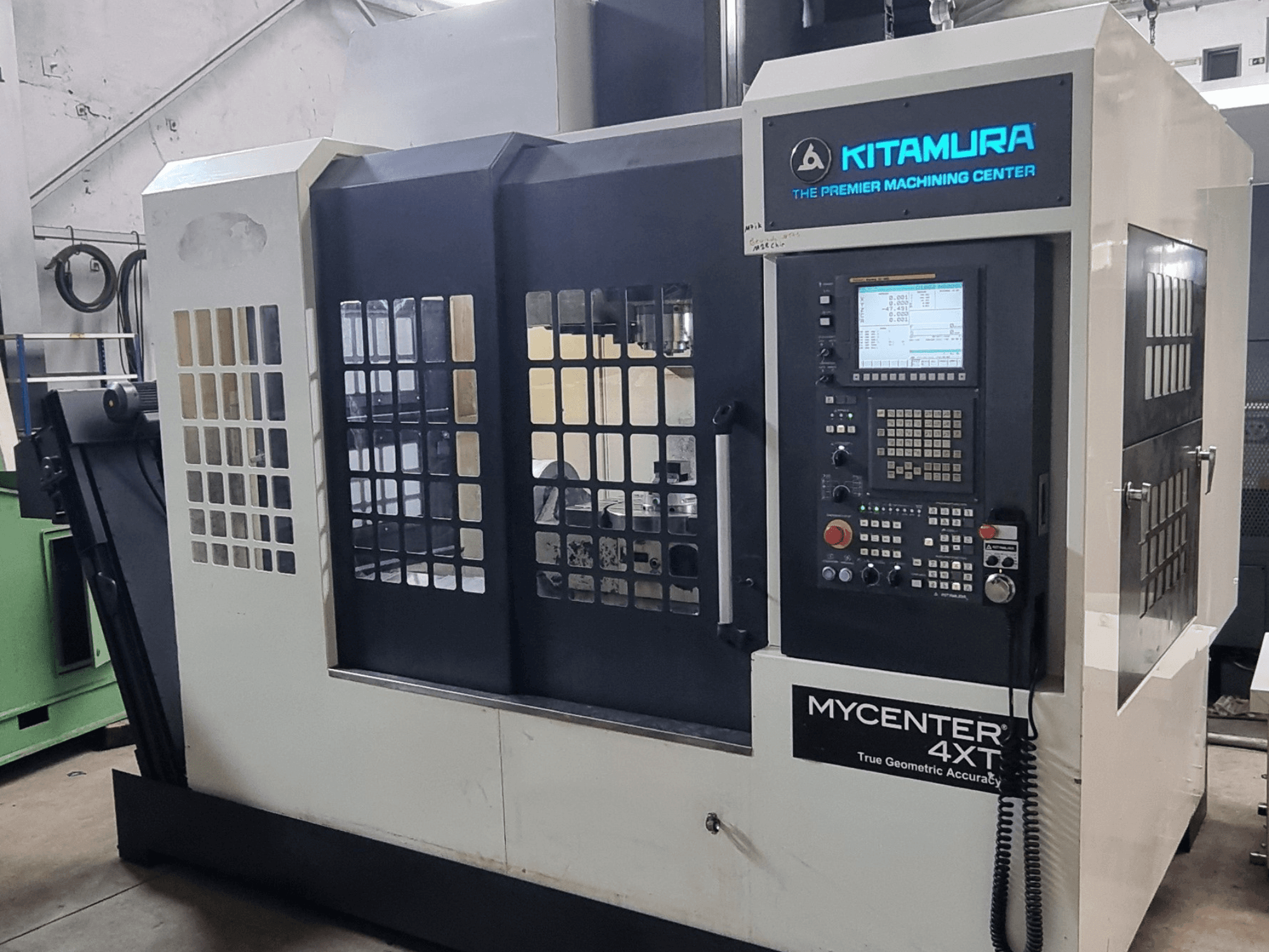 Альтернативный текст: Вертикальный обрабатывающий центр Kitamura MYCENTER 4XT, вид спереди, на котором показаны панель управления и детали станка в условиях цеха.